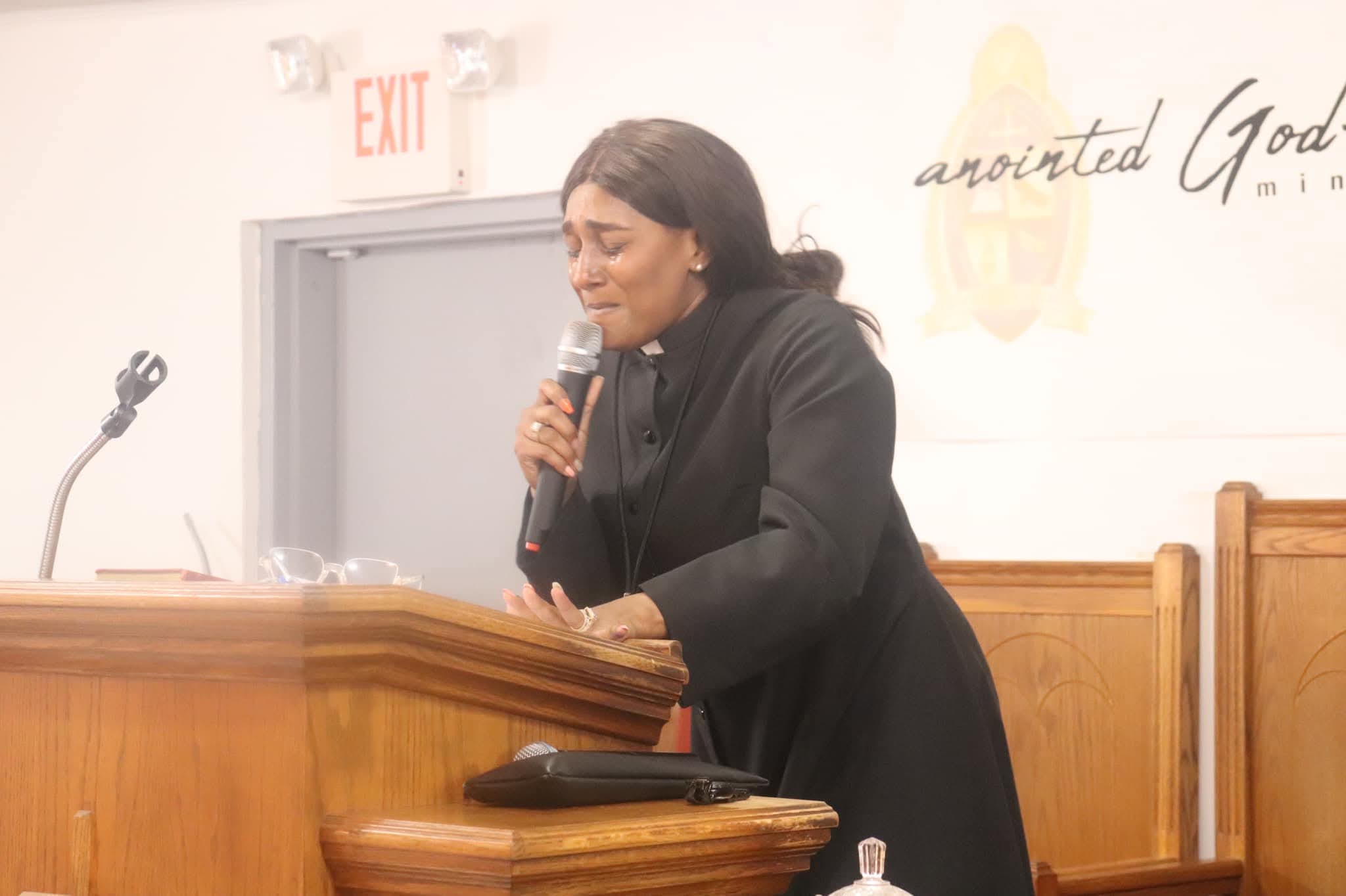 Woman speaking into microphone at podium in a room with wooden furniture and a white wall.