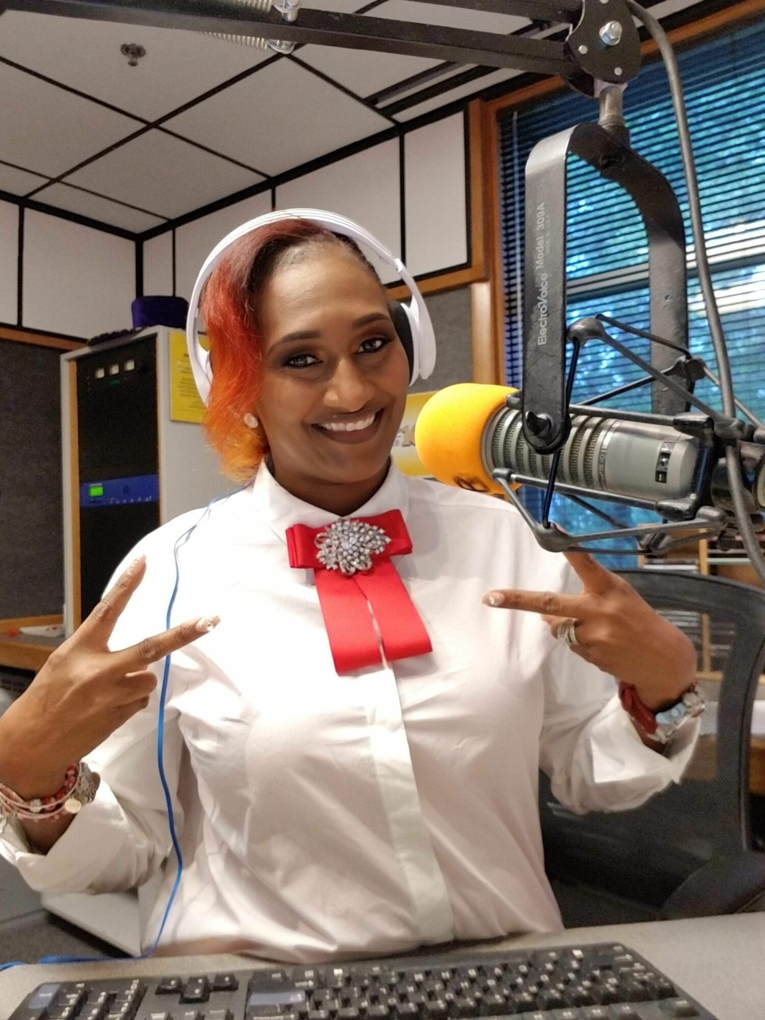 Woman with orange hair in a white outfit, smiling, pointing at a microphone in a recording studio.