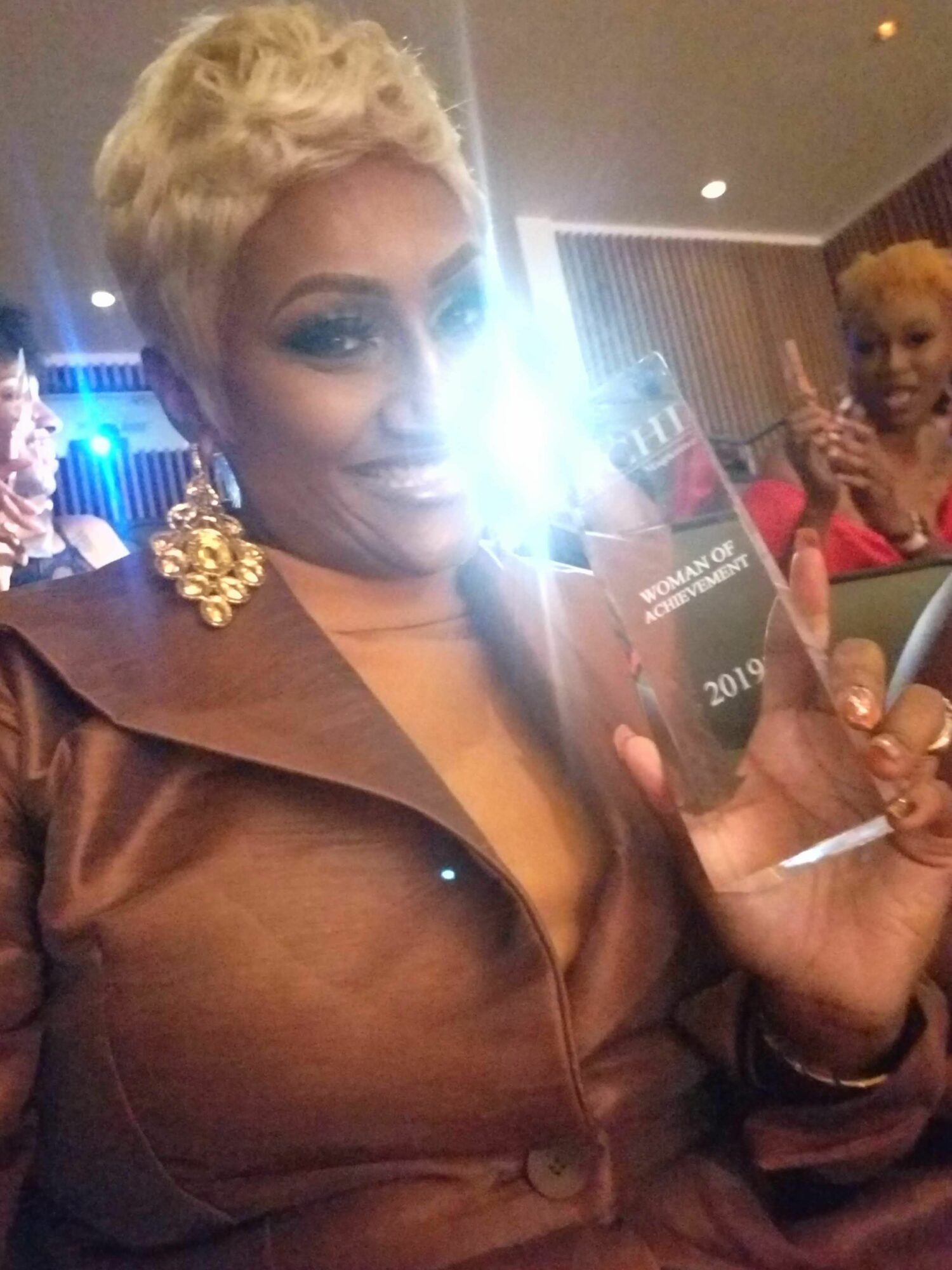 Woman with short blonde hair holding a phone with a bright flash, wearing a brown jacket, in an indoor setting.