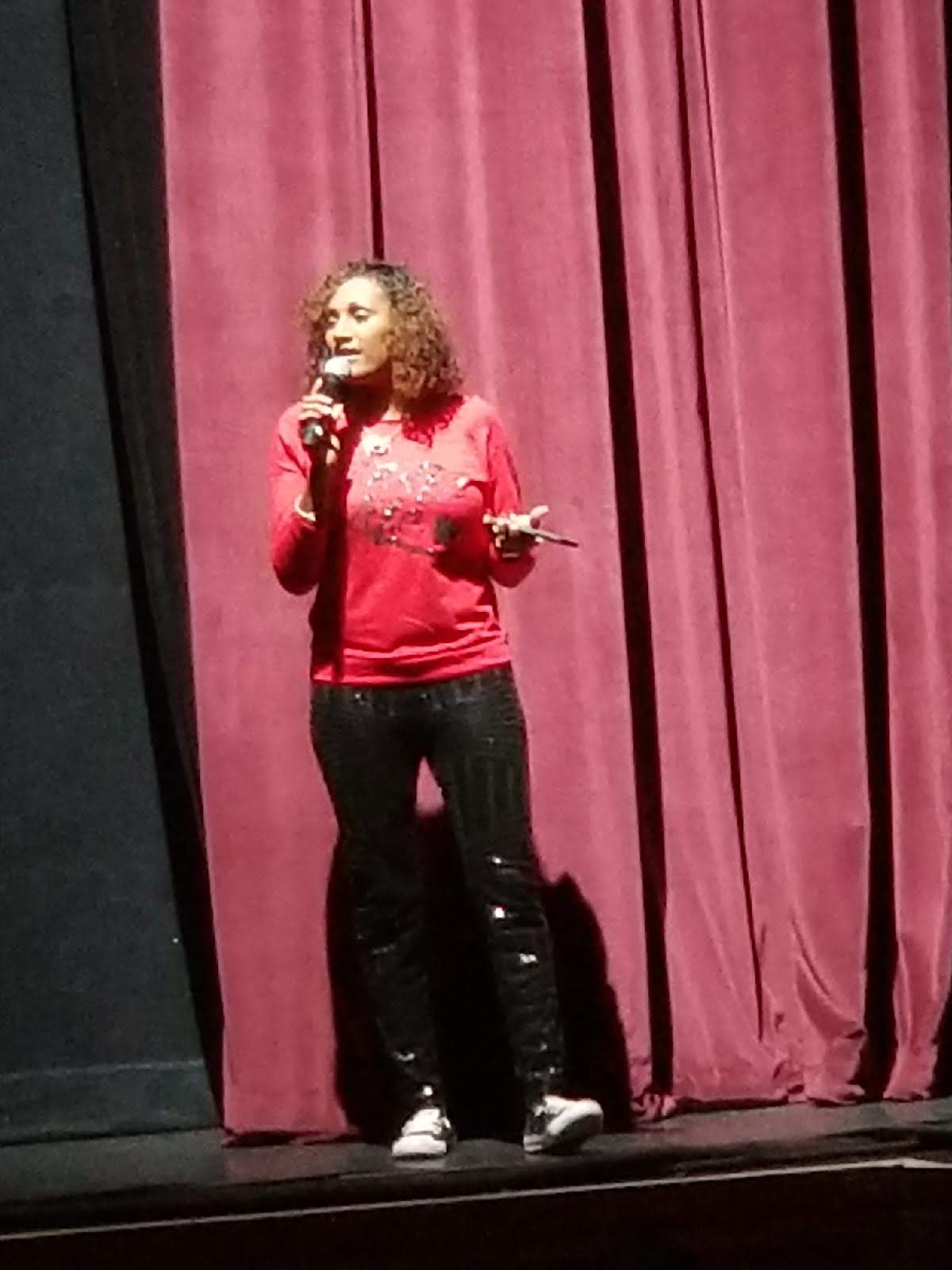 Woman standing on stage with a microphone in front of a pink curtain, holding a paper, speaking to an audience.