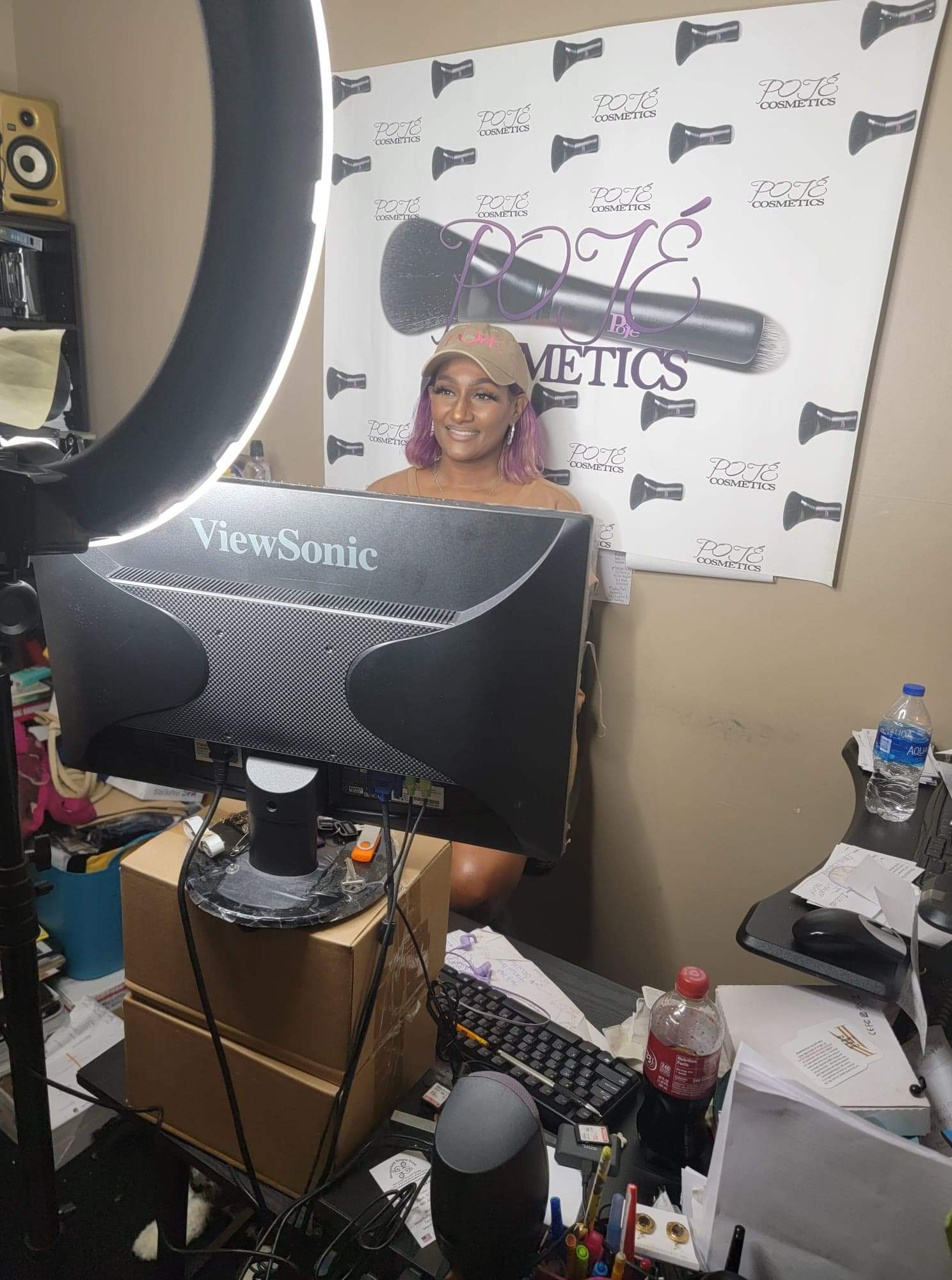Person with purple hair smiling in front of a cosmetics banner, surrounded by computer equipment and bottles.