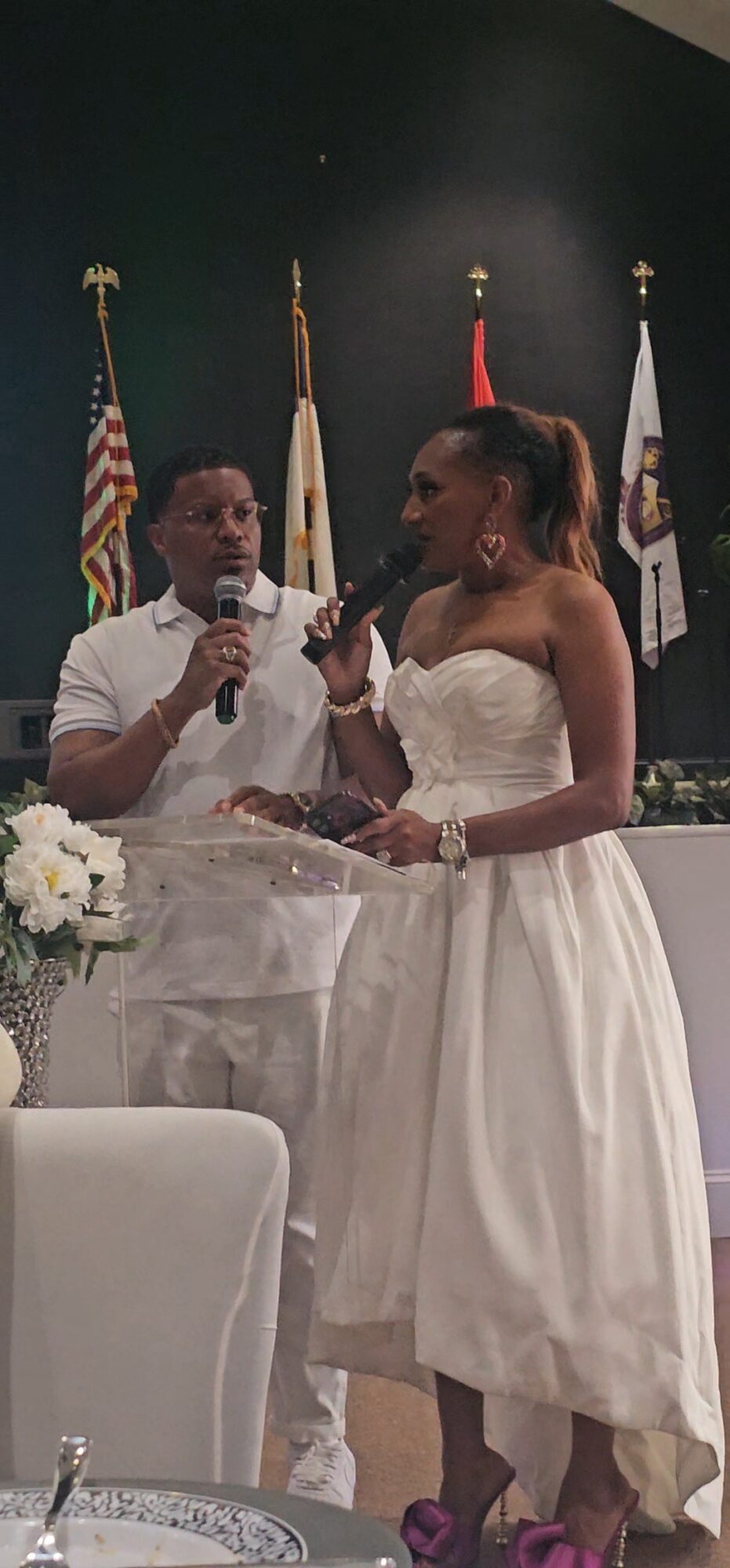 Two women in wedding dresses stand together, one holding a microphone, with flags in the background.