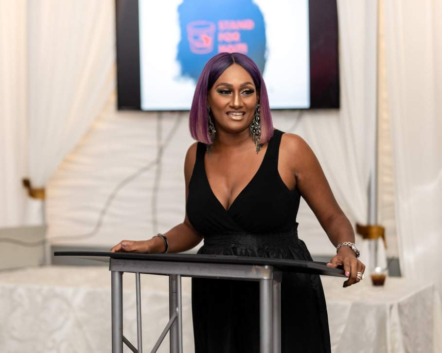 Woman with purple hair in a black dress standing behind a table, smiling, in a room with white curtains and a screen.
