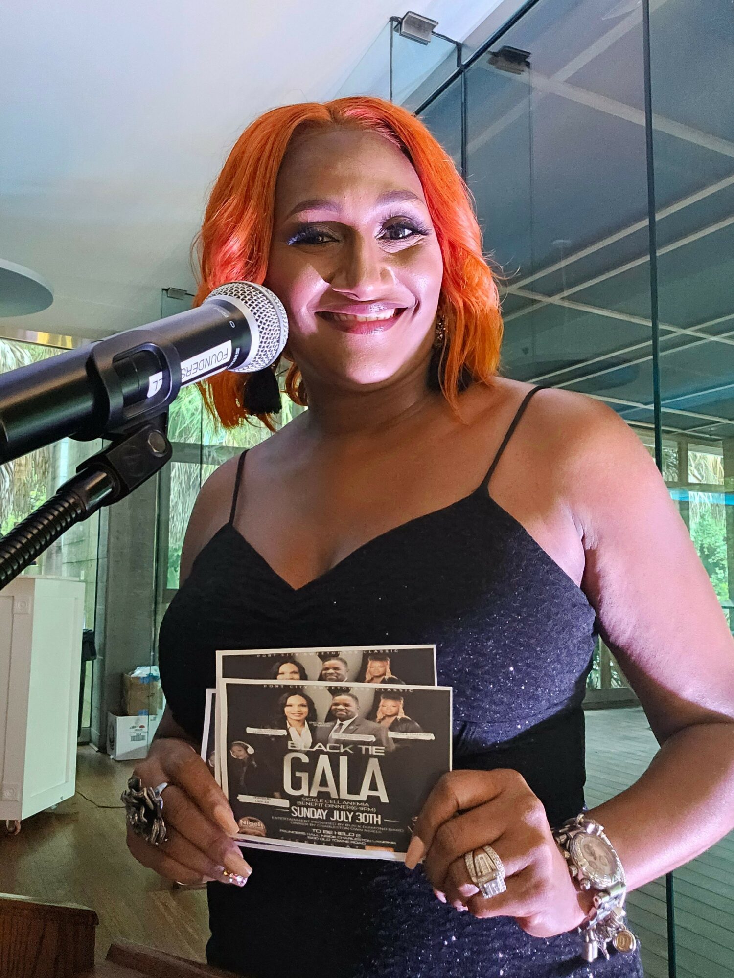 Woman with orange hair holding a book and speaking into a microphone, standing indoors near glass windows.