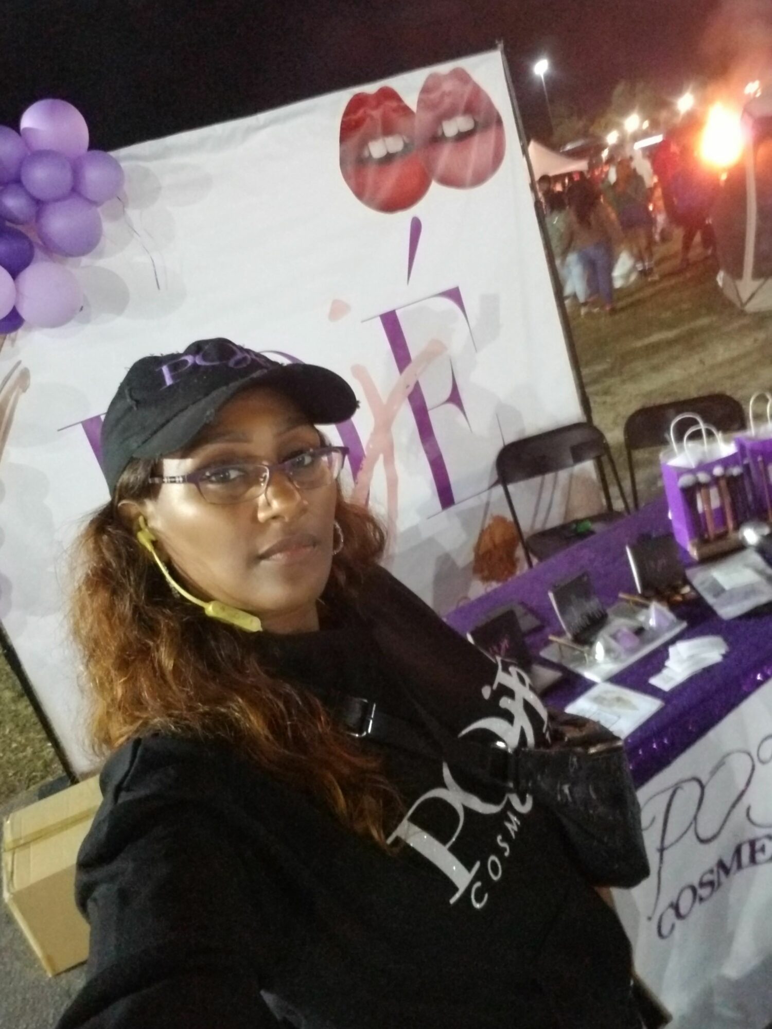 Woman with glasses and curly hair wearing a black cap and jacket, standing at a table with purple balloons and decorations.