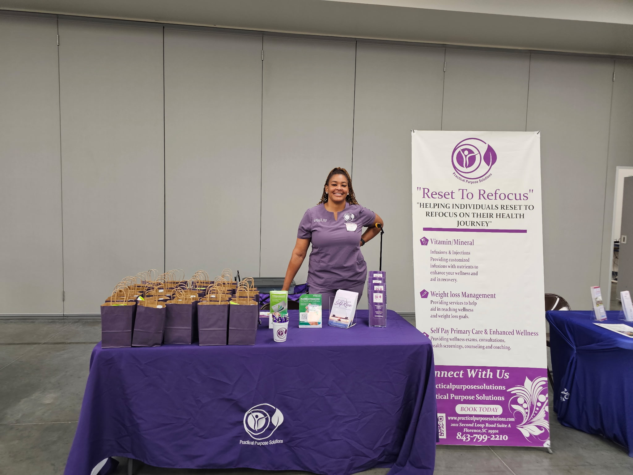 Woman standing behind a table with health products, purple tablecloth, and a vertical banner, in a room with gray walls.