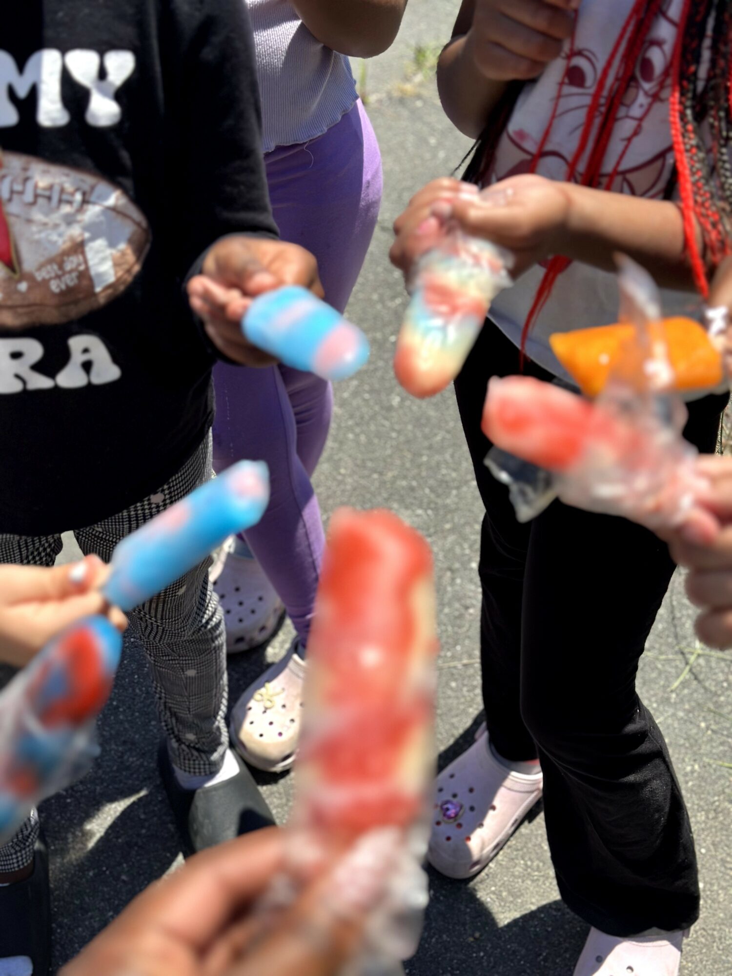 Children holding colorful ice pops outdoors, some with hands visible, wearing casual clothing and sneakers.