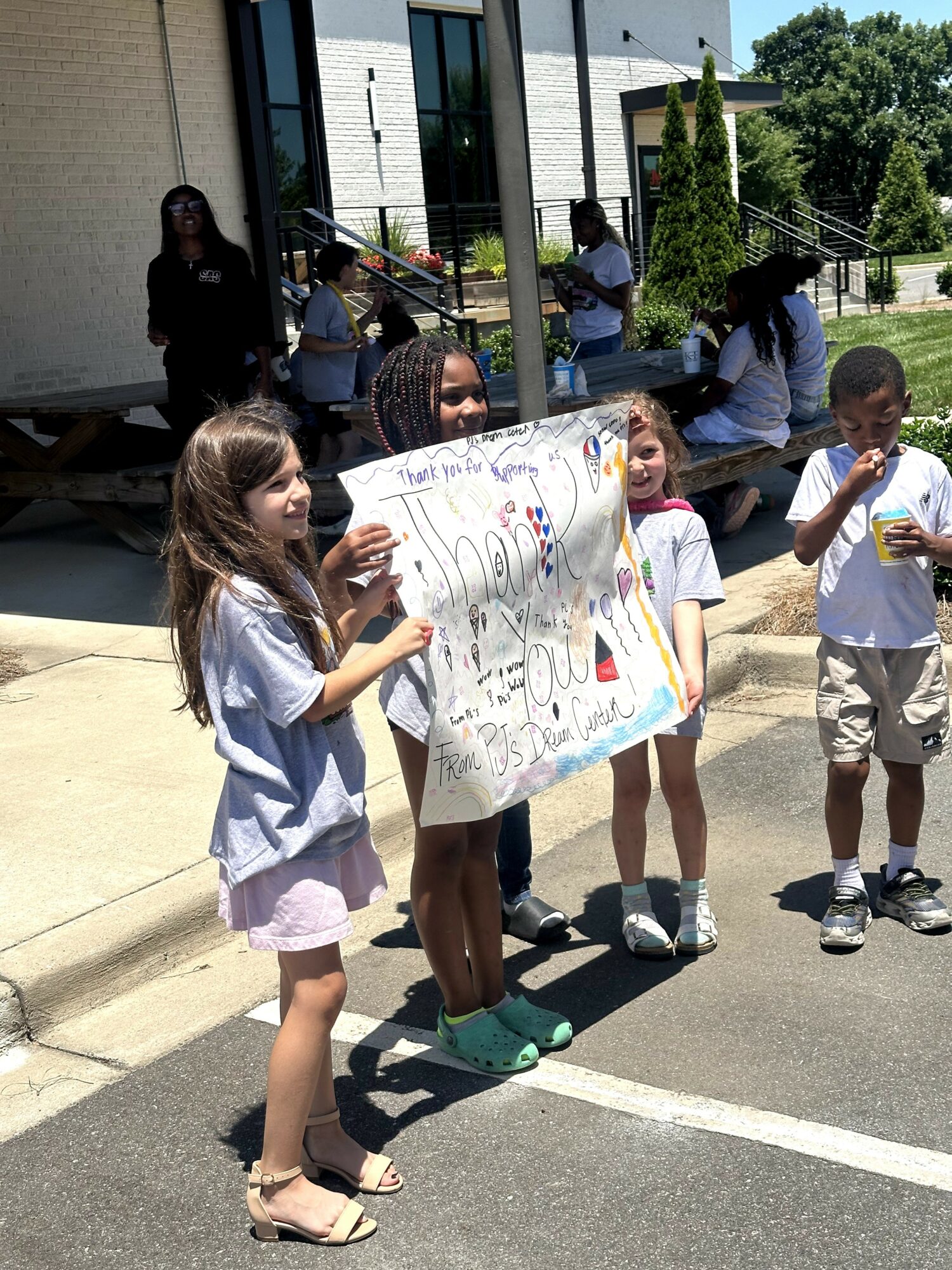 Group of children holding a colorful poster outside a building, with adults nearby and trees in the background.