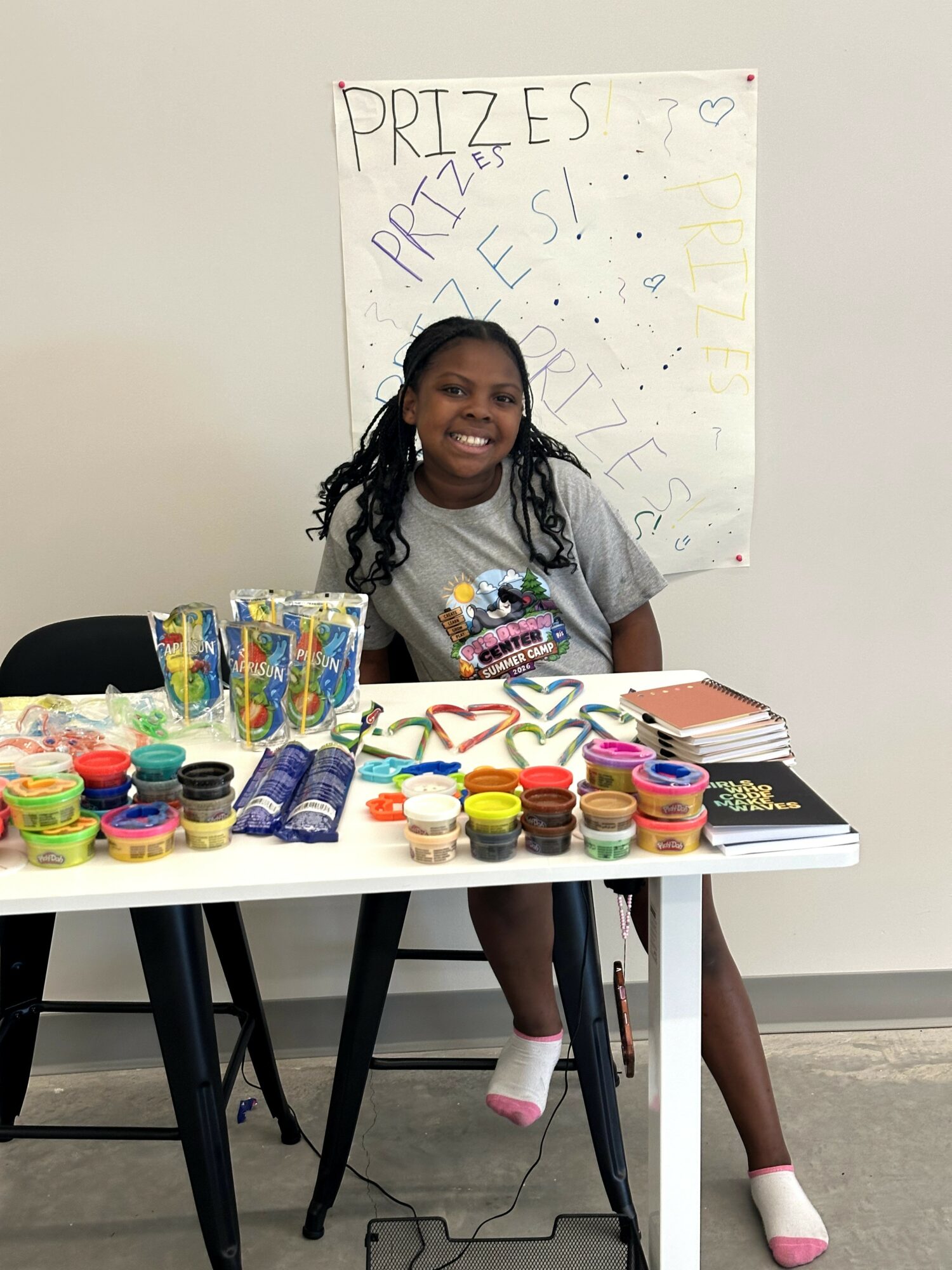 Girl sitting at a table with colorful prizes and a sign behind her, smiling, in a room with a white wall.