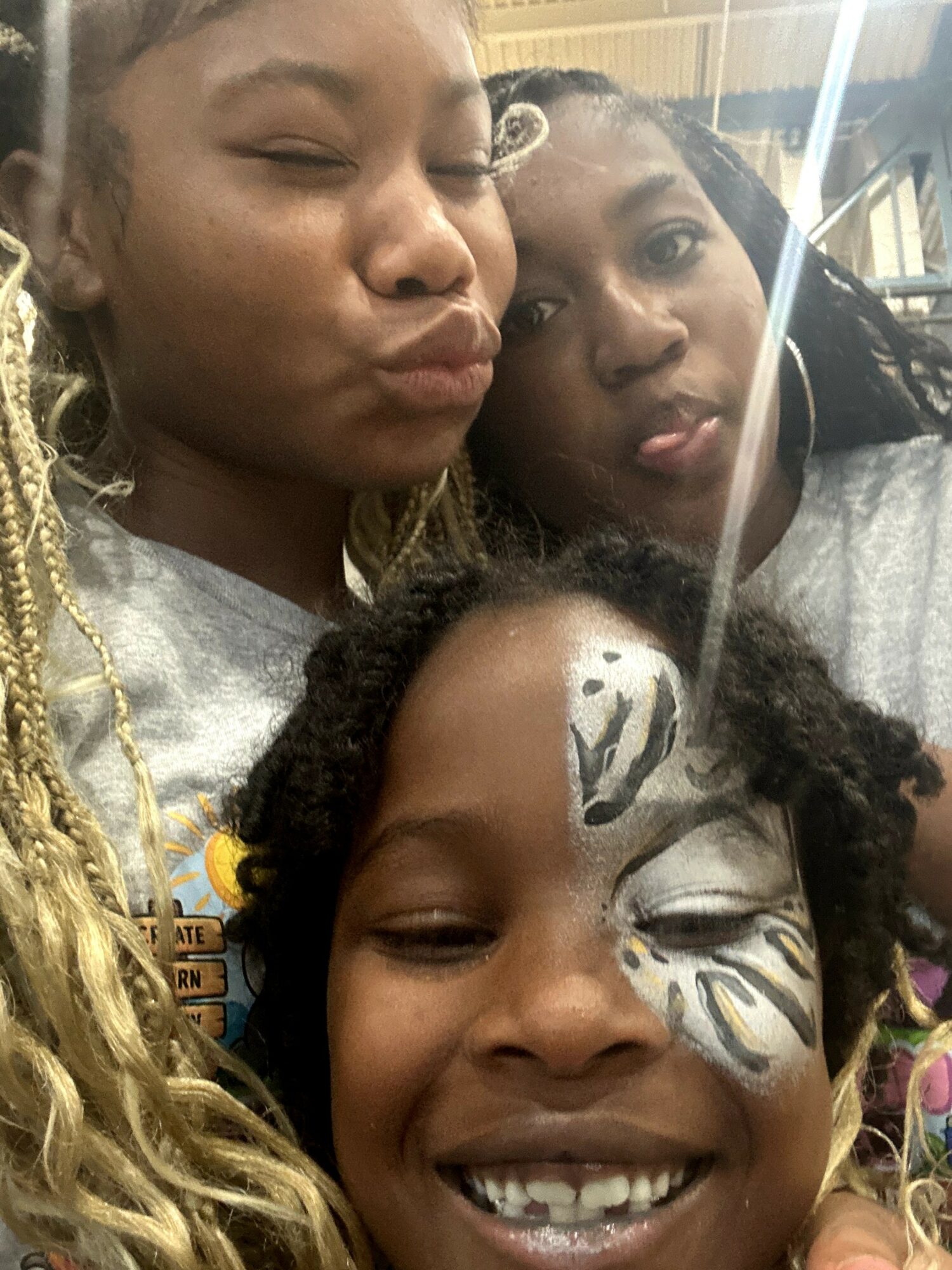 Three smiling children, one with face paint, close together indoors, with a bright background and reflections.