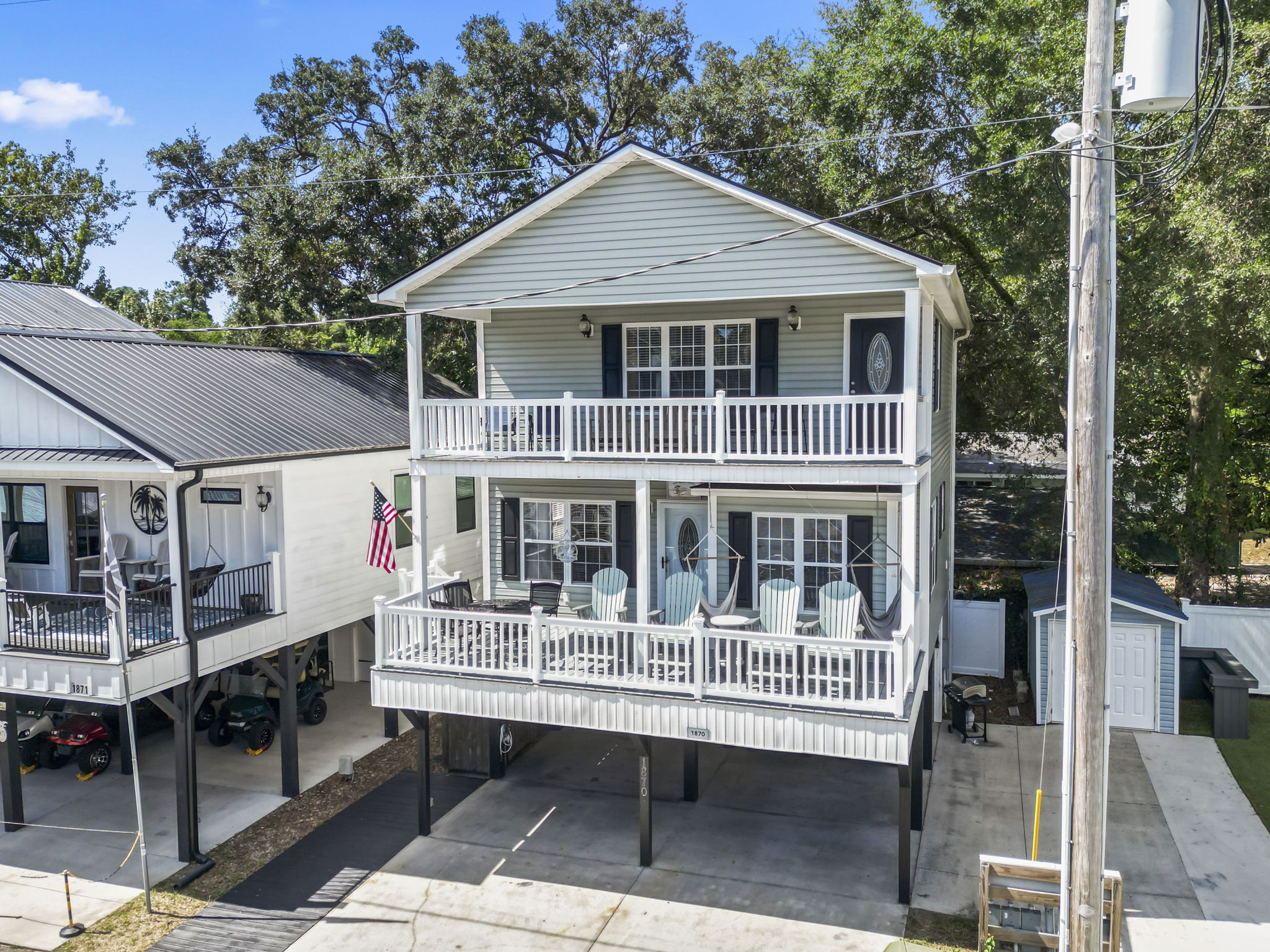 Three-story house with balconies, white railings, and large windows, elevated on stilts, with a driveway and trees in background.