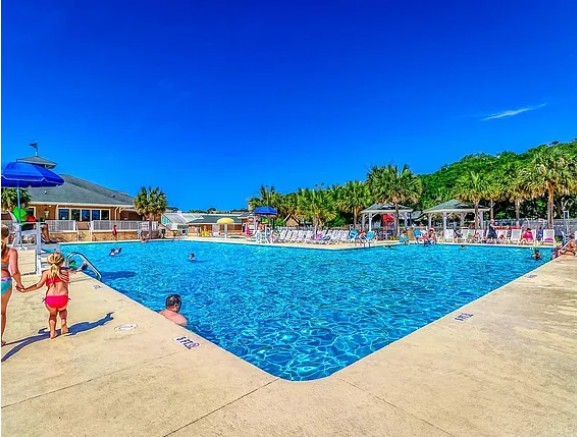 People swimming and relaxing at an outdoor pool with blue sky and trees in the background.