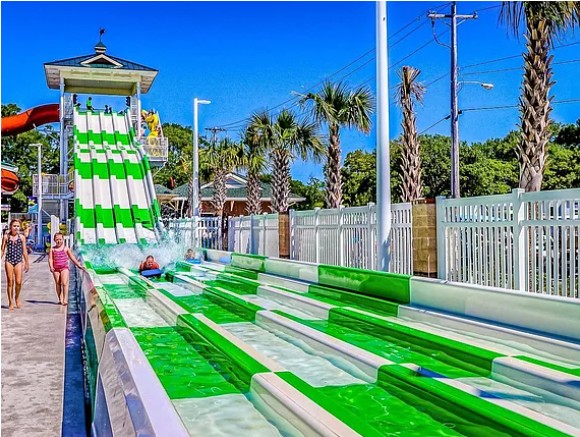 People at a water slide with green and white lanes, palm trees, and a white fence in the background.