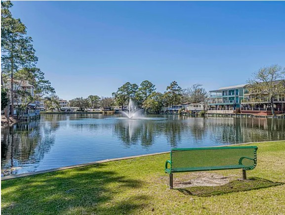 A park with a lake, fountain, green bench, trees, and buildings in the background under a clear blue sky.