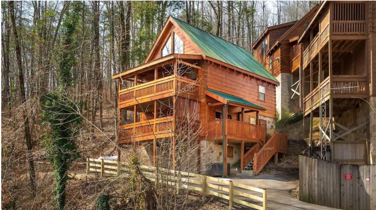 Wooden house with green roof on a wooded hillside, surrounded by trees and a wooden fence, with stairs leading up to it.
