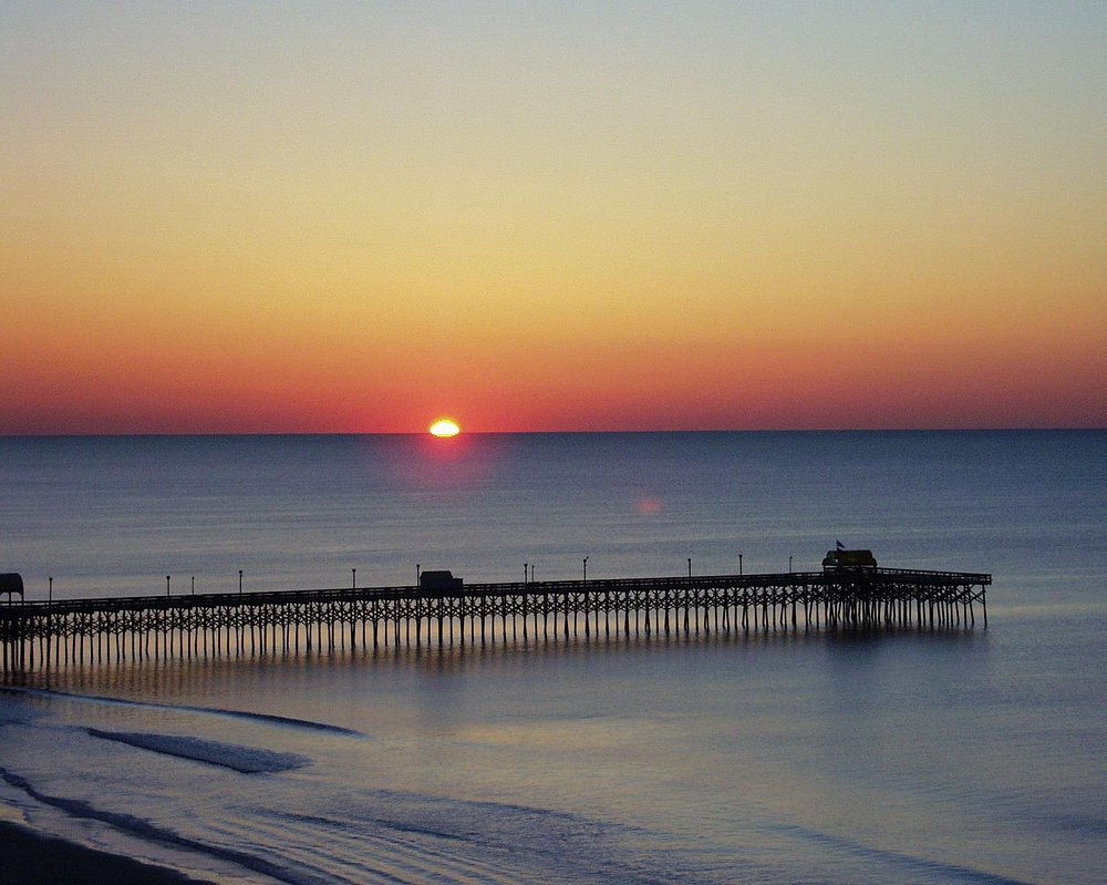 Sunset over the ocean with a pier extending into the water, calm waves, and a colorful sky.