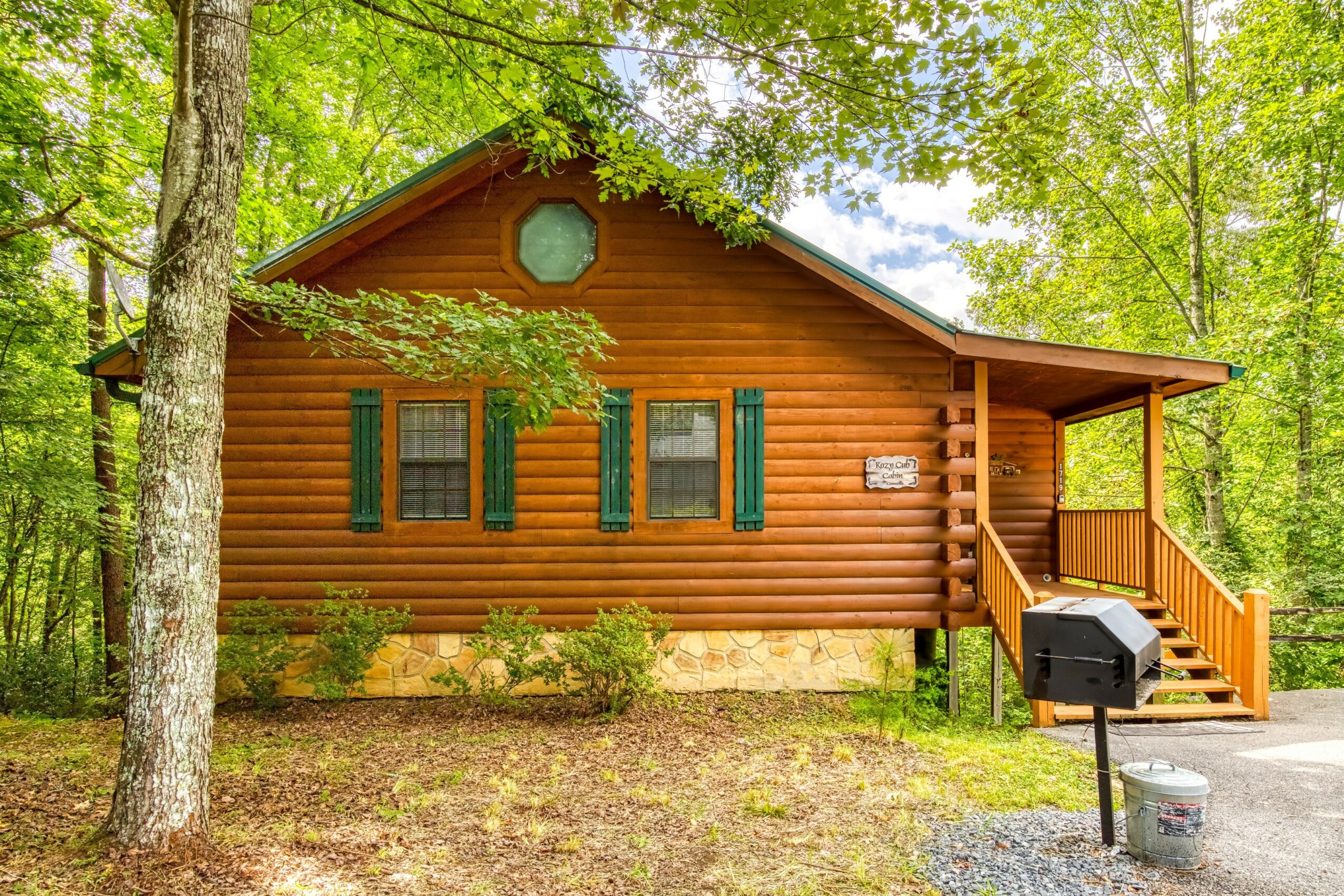 Wooden cabin with green shutters, surrounded by trees, with a porch and a grill outside, in a forest setting.
