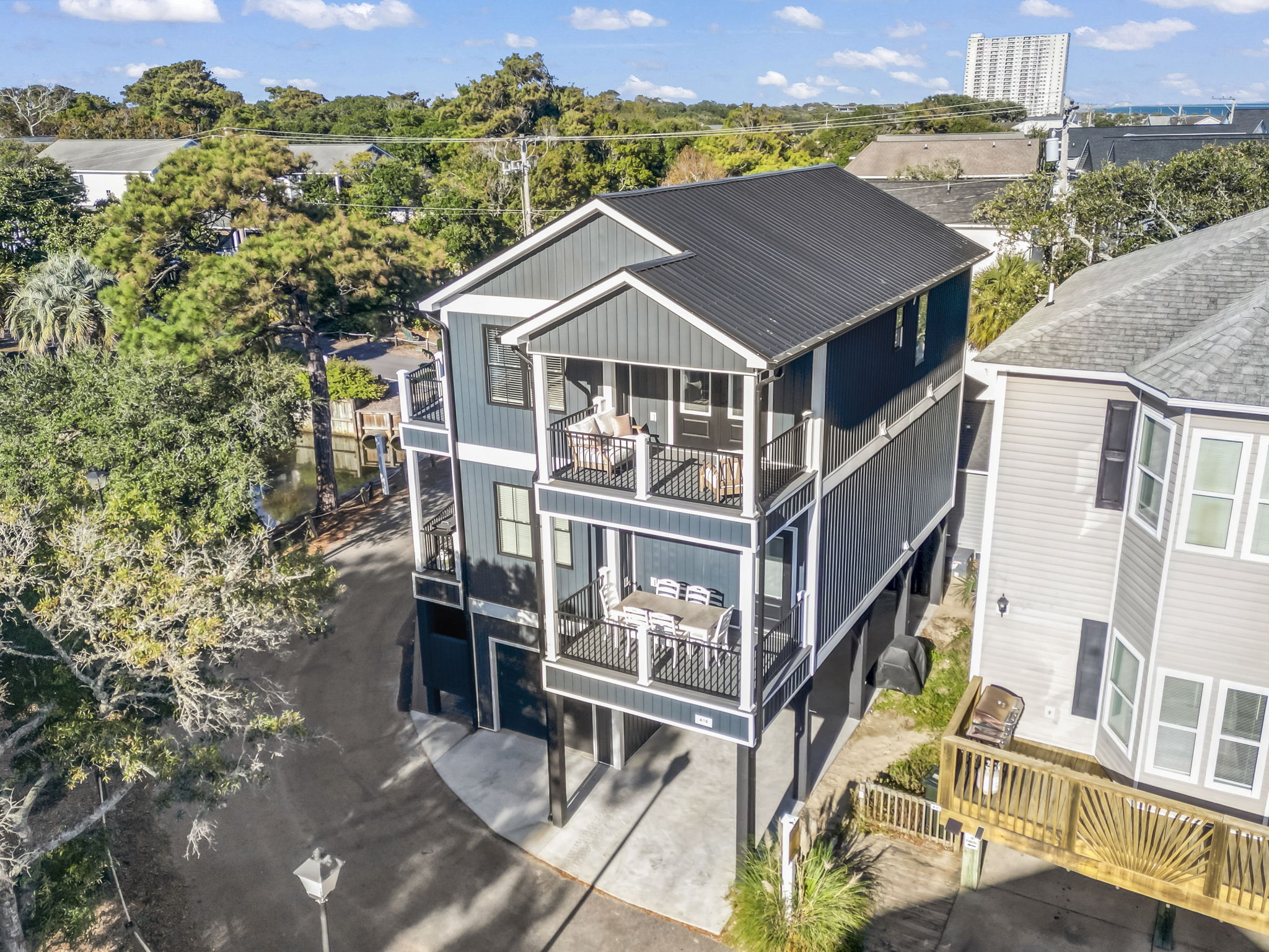 Three-story house with balconies, surrounded by trees and neighboring houses, under a blue sky with clouds.