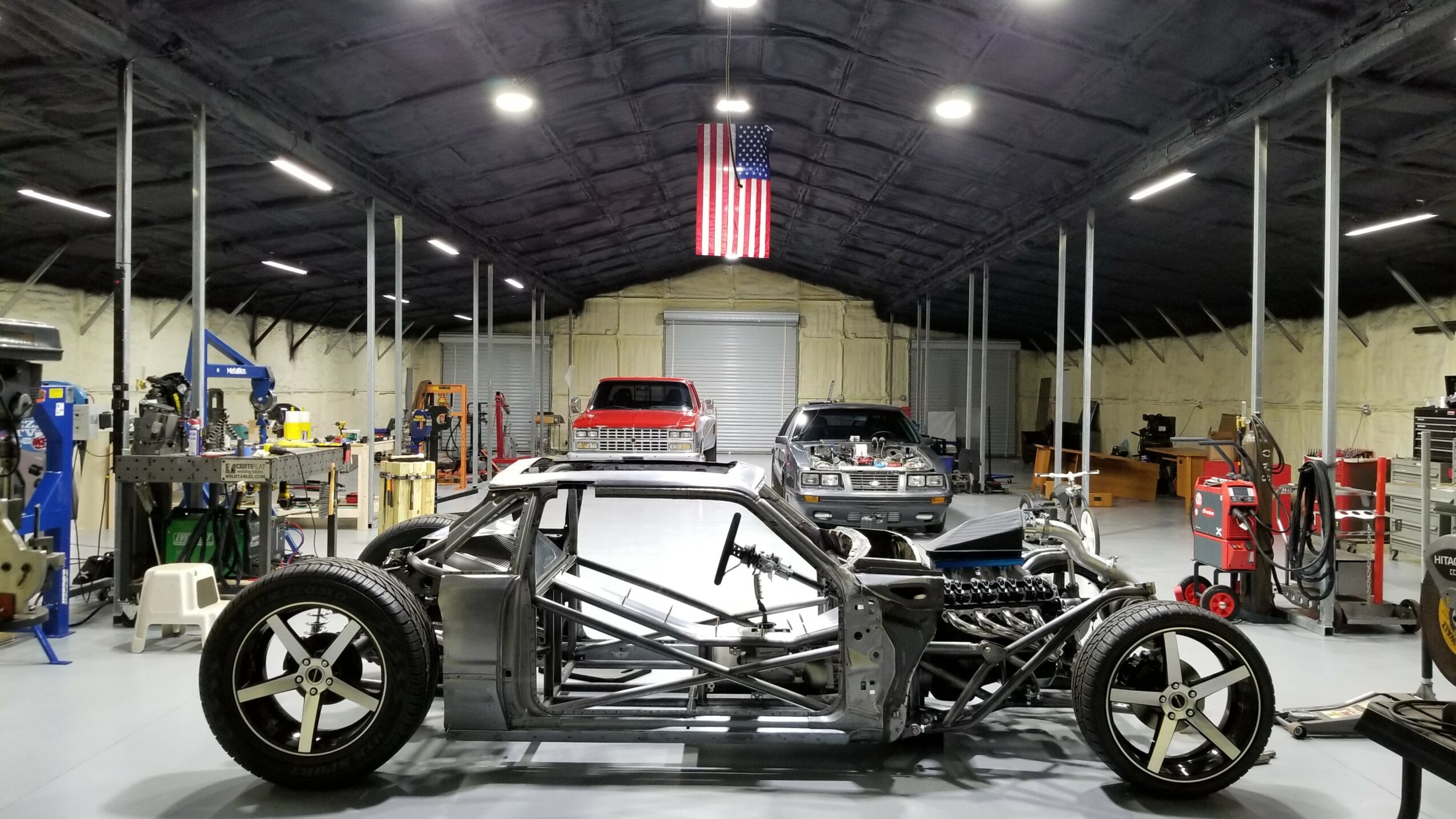 Empty automotive workshop with a partially assembled race car in the center, surrounded by tools and equipment.