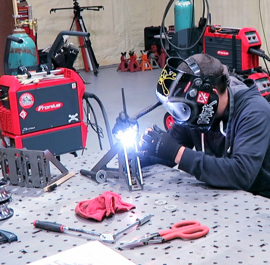 Person welding metal parts at a workbench with welding helmet, tools, and equipment in workshop.