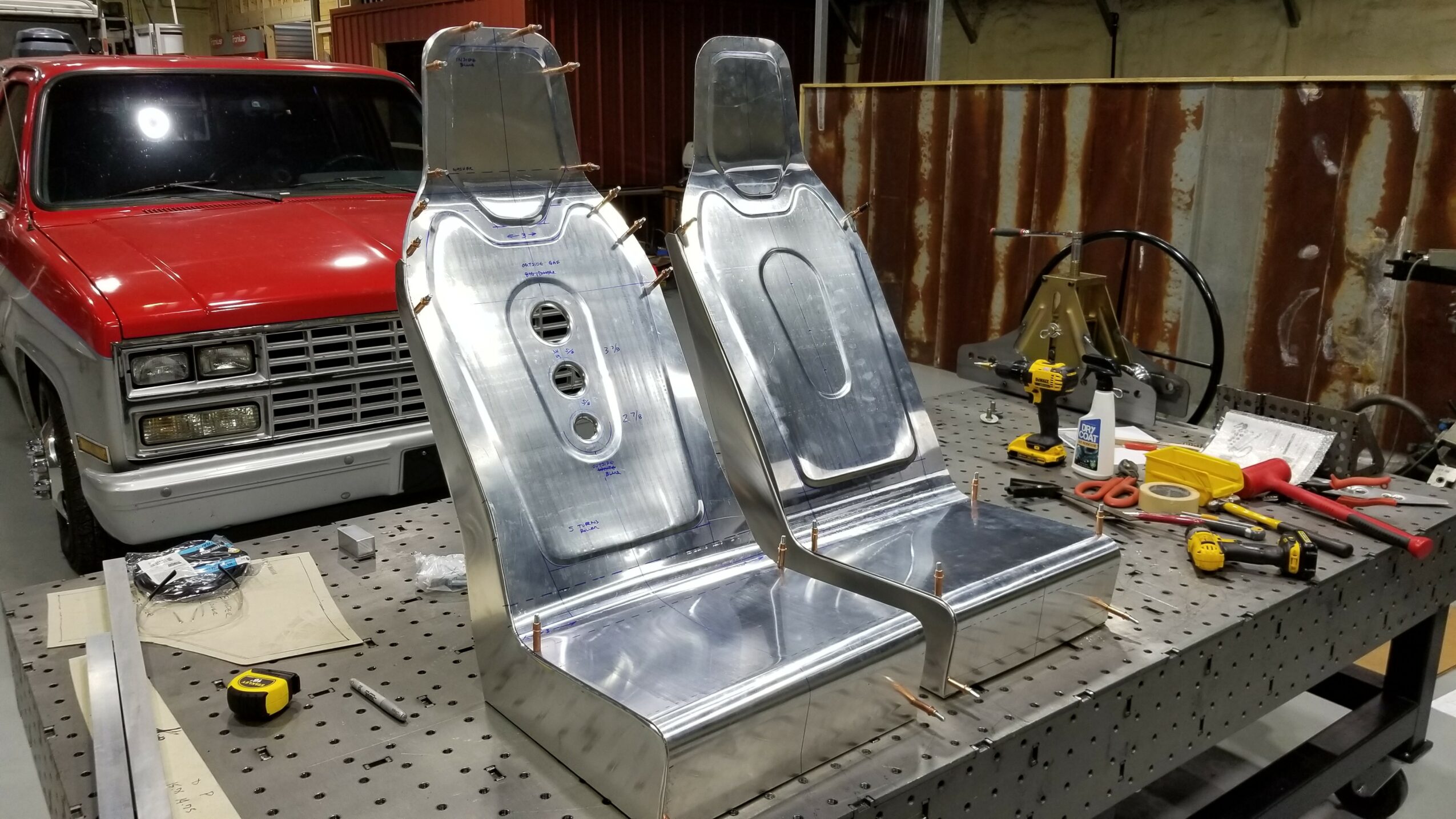 Two metallic car seats on a workbench with tools and equipment nearby, in a workshop setting.