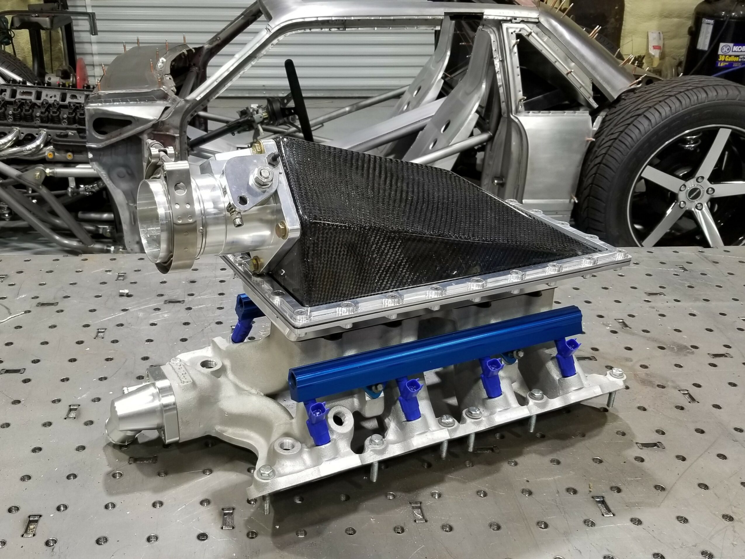 Automotive engine component with a black heat shield and blue mounting parts, placed on a workbench in a garage.