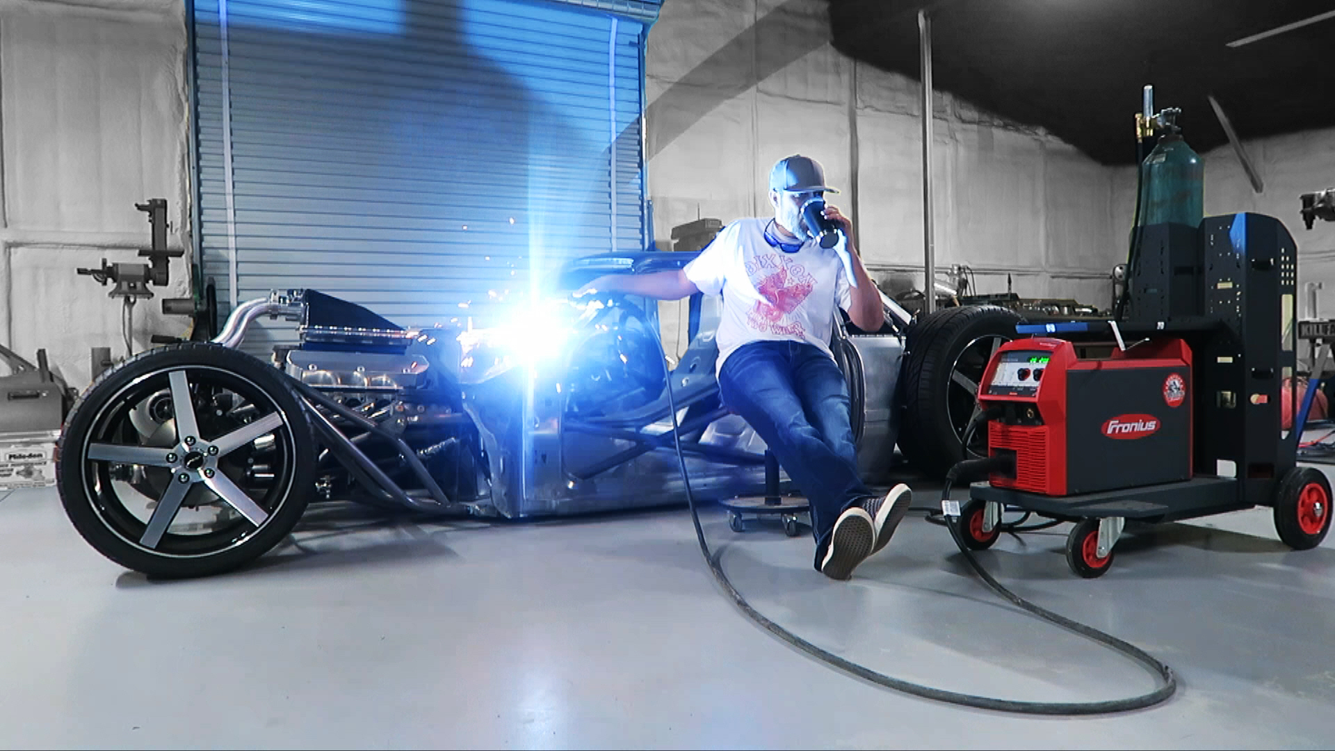 Person welding a vehicle frame in a workshop with welding equipment nearby.