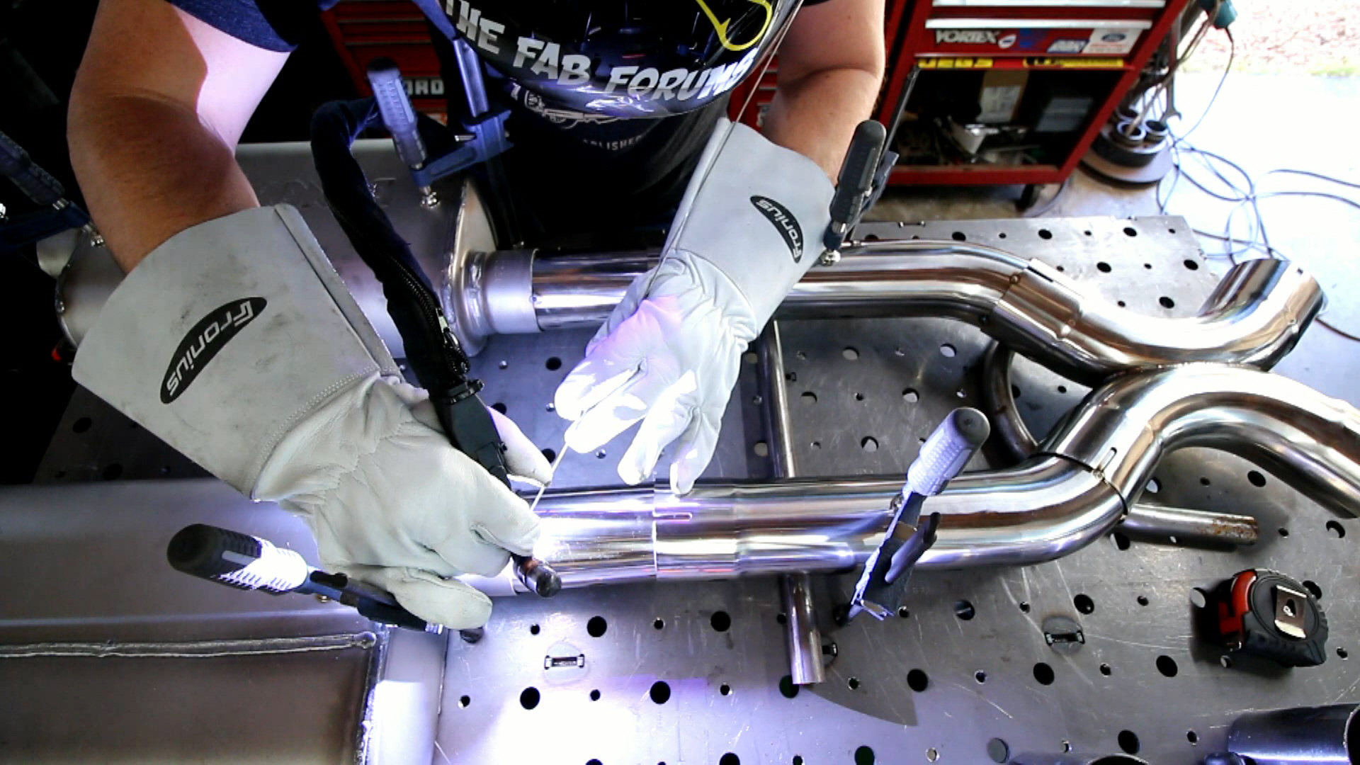 Person welding metal pipes on a workbench, wearing gloves and protective gear, with tools and equipment nearby.