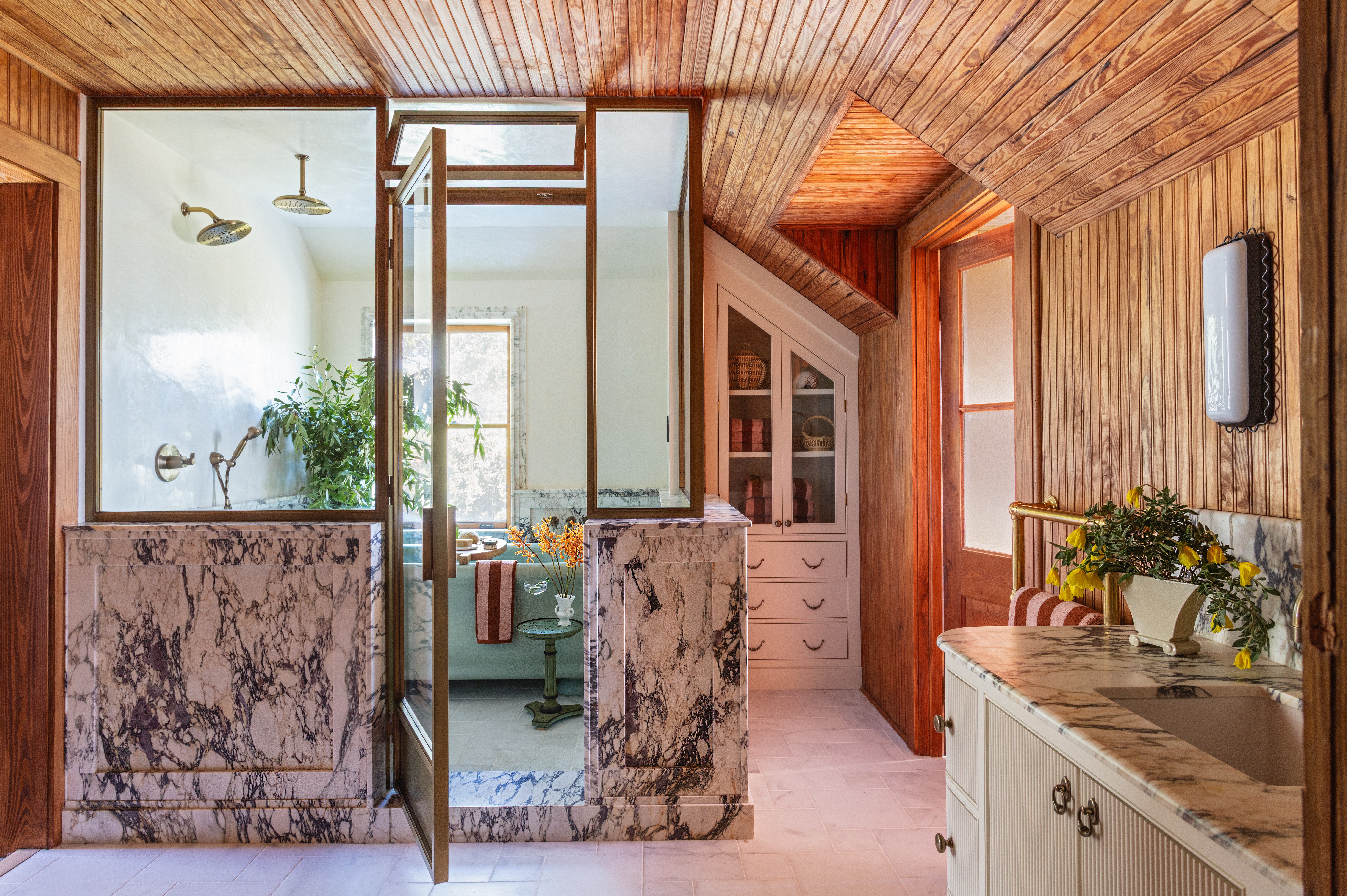 View of a kitchen with wooden walls and ceiling, a marble counter, and a window with a plant. Bright natural light.