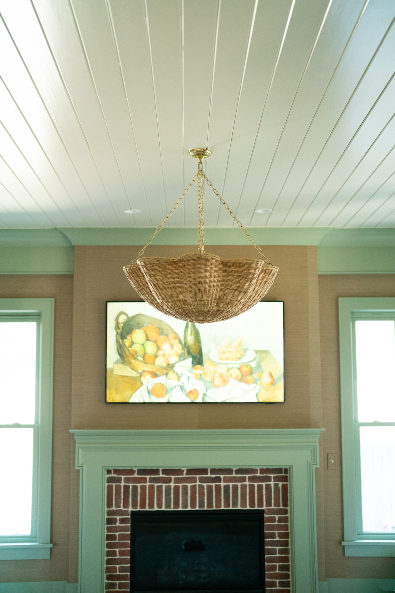 Chandelier hanging above a fireplace with a painting on the wall, two windows on either side, and a white ceiling.