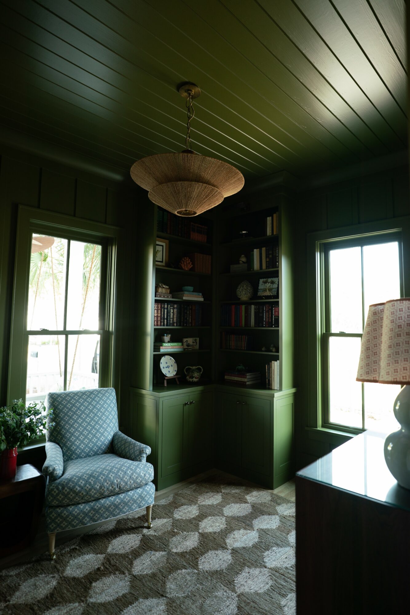 Room with green walls, ceiling, and built-in bookshelf, two windows, patterned armchair, and a lamp, with a patterned carpet.