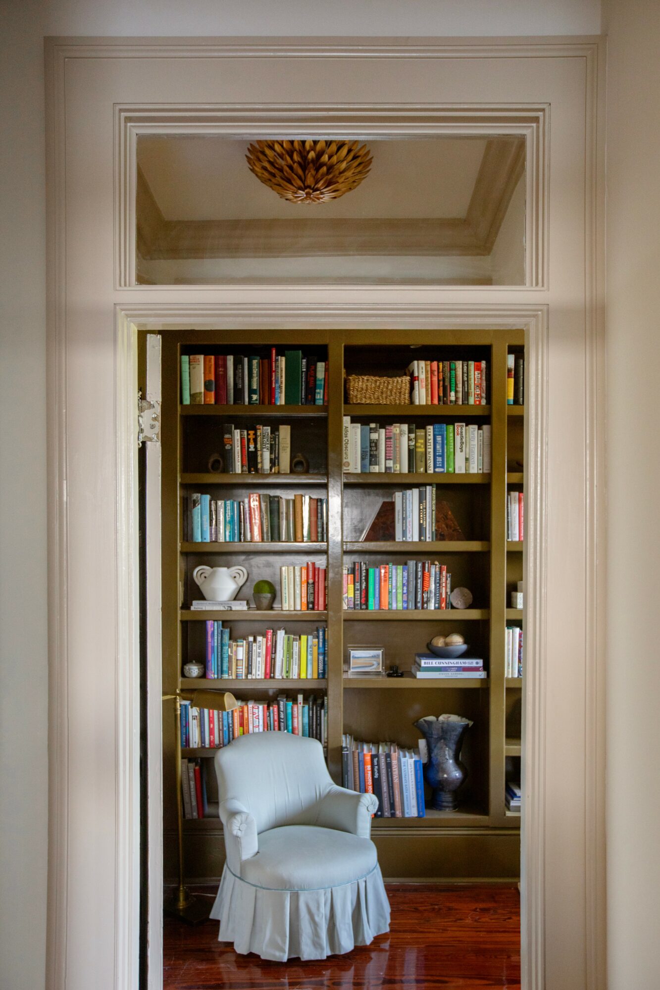 Bookshelf filled with books and decorative items, white chair with a skirt in front, ceiling with decorative element, door frame.