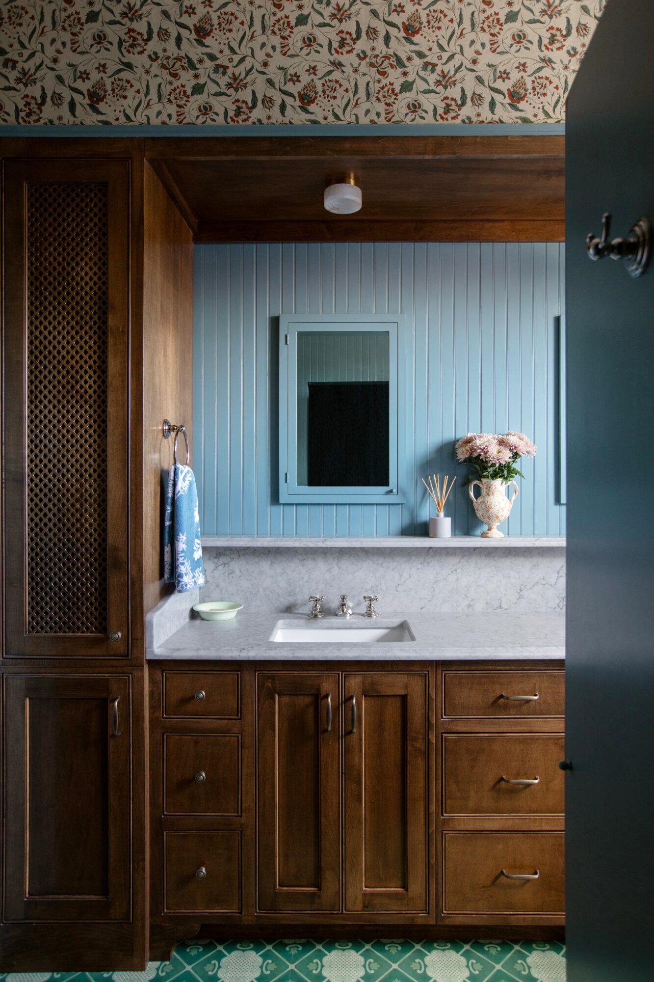 Bathroom mirror above sink, wooden cabinets, vase with pink flowers, blue wall, floral ceiling, partially open door