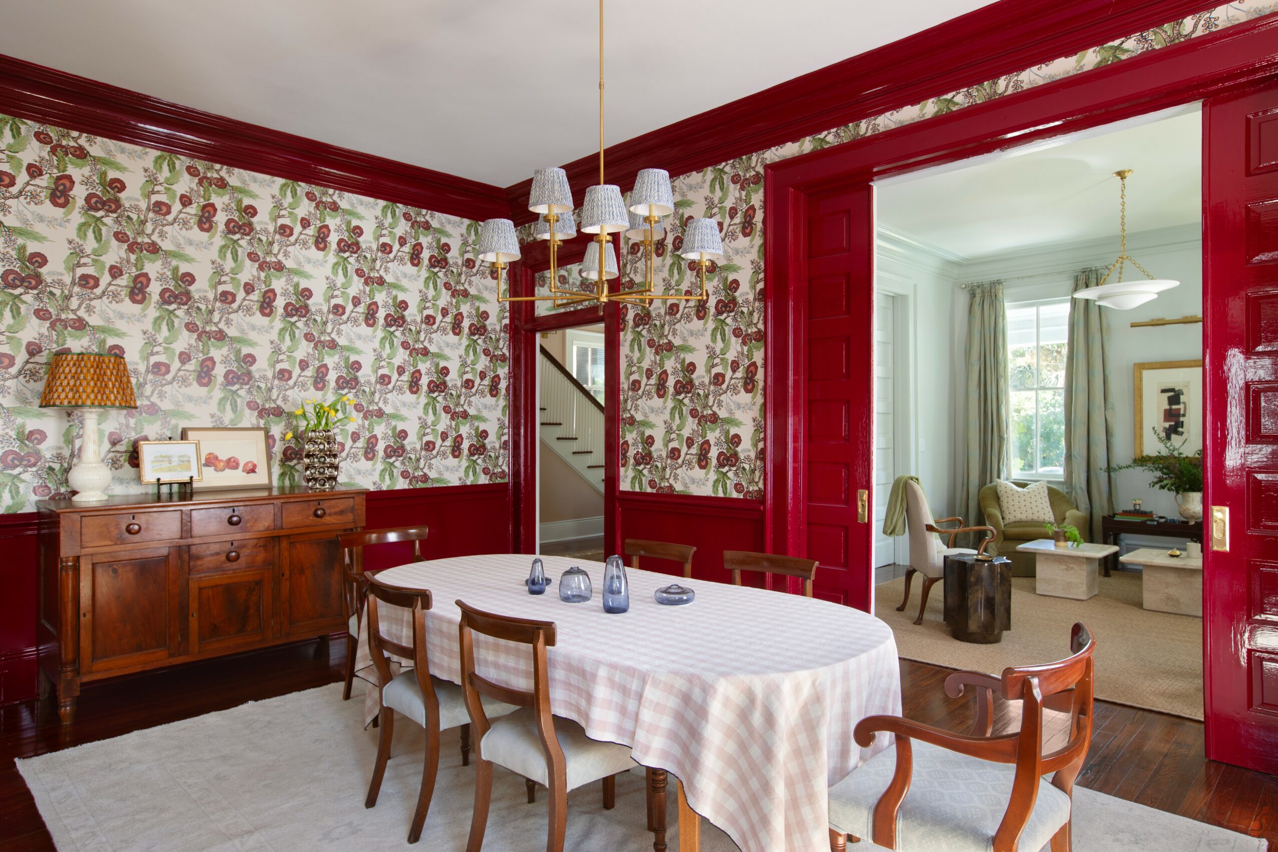 Dining room with floral wallpaper, wooden sideboard, white tablecloth, and chairs, connected to a bright living room through red doorway.