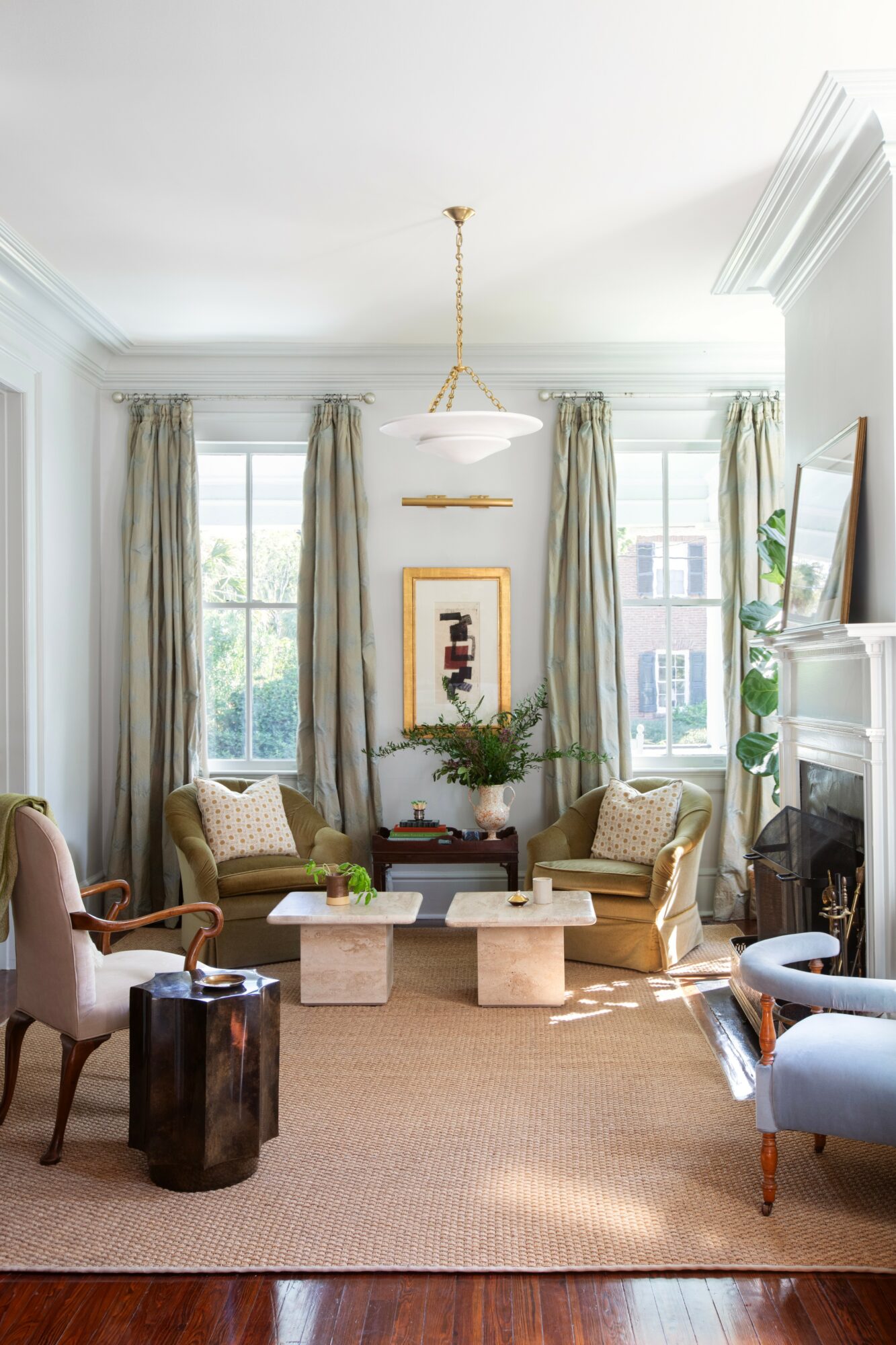 Living room with large windows, curtains, two armchairs, a sofa, a fireplace, and a chandelier, decorated in neutral tones.
