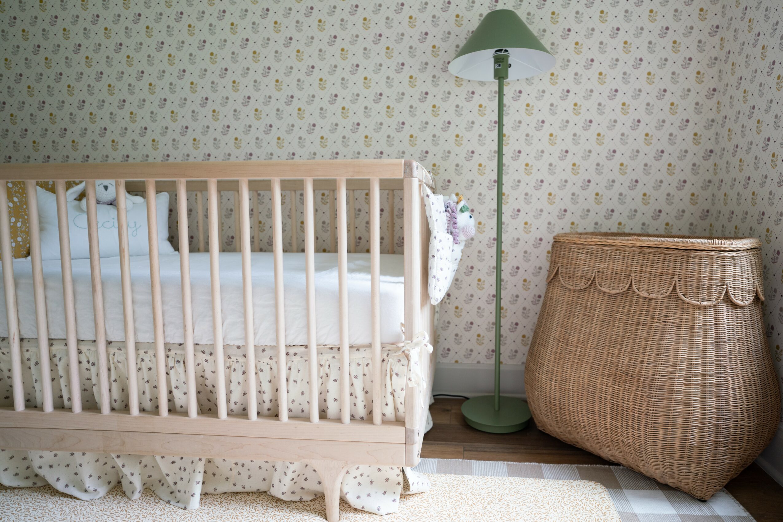 Baby crib with bedding, a tall green floor lamp, and a woven basket in a room with floral wallpaper.