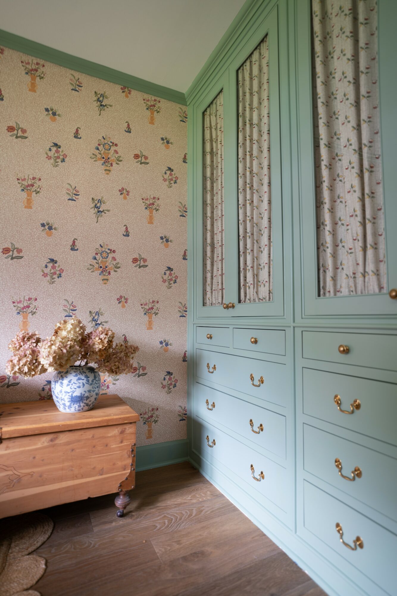 Corner of a room with floral wallpaper, a wooden chest, and a light green cabinet with drawers and patterned curtains.