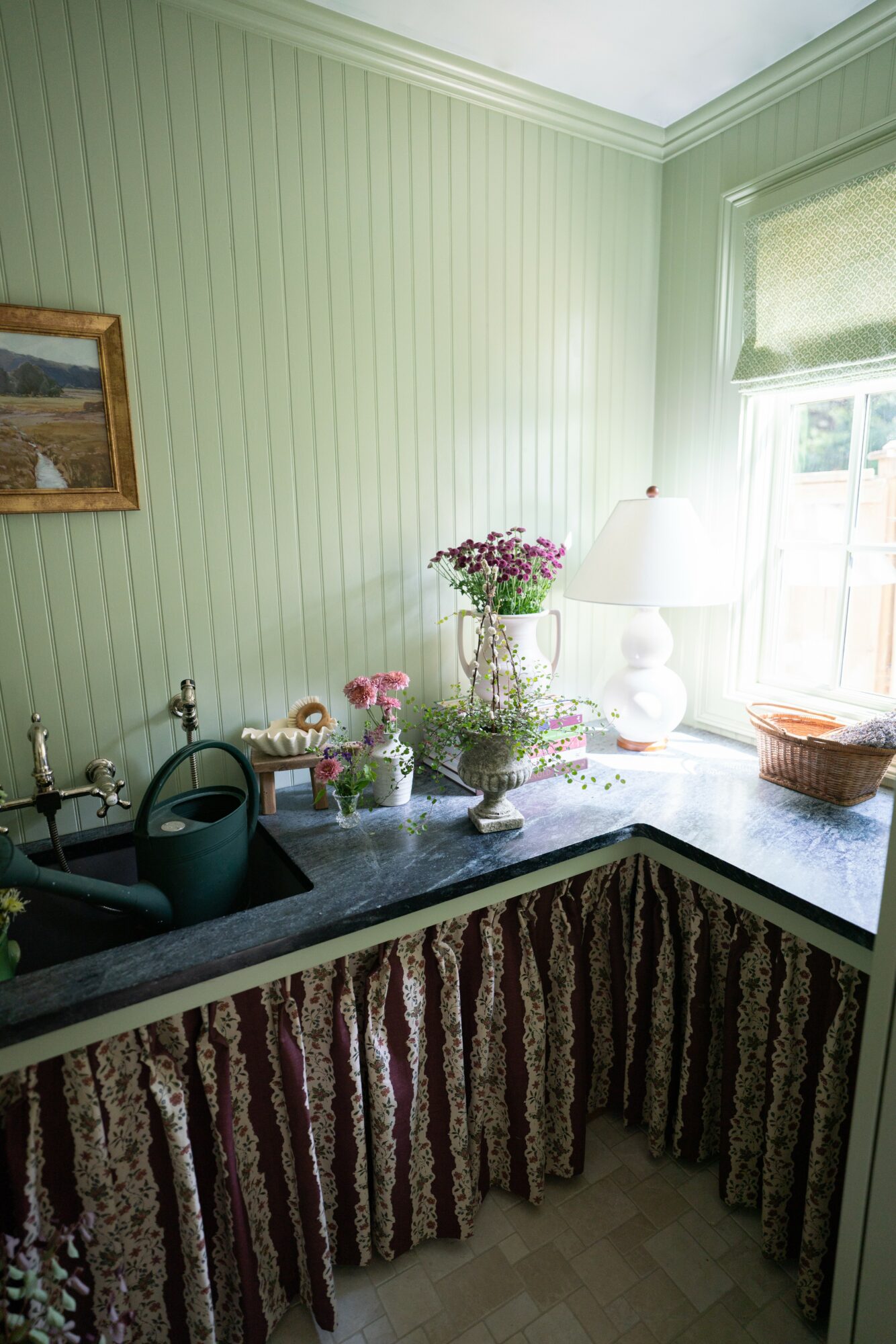 Corner of a room with a window, green paneled walls, a countertop with flowers, a lamp, and a basket.