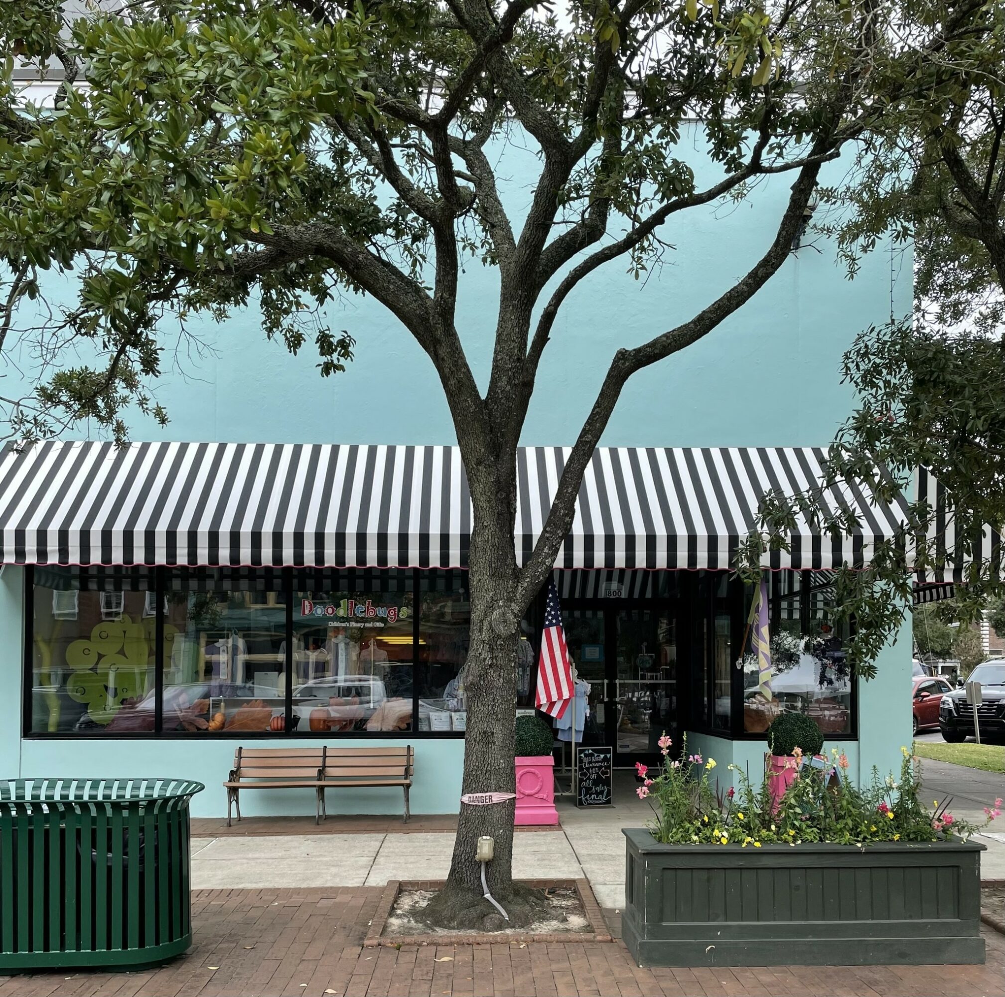 Tree in front of a building with striped awning, benches, and flower planters, on a sidewalk.