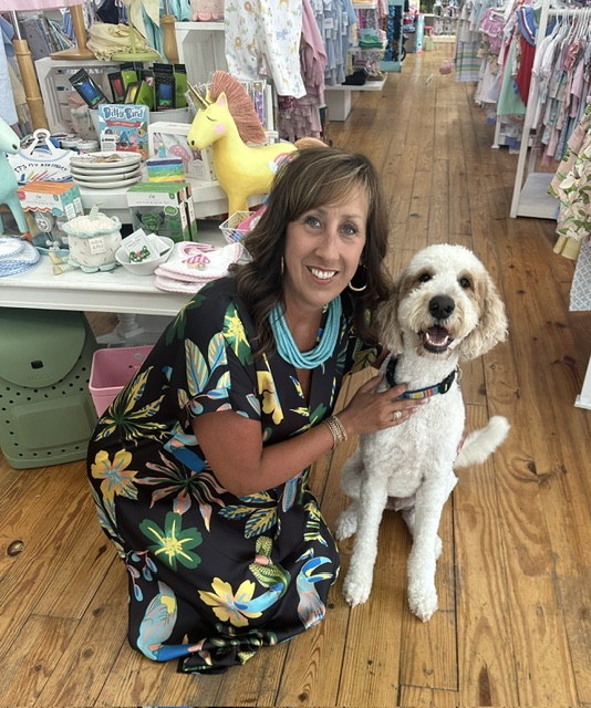 Woman kneeling next to a white dog inside a store with shelves of household items and clothing in the background.