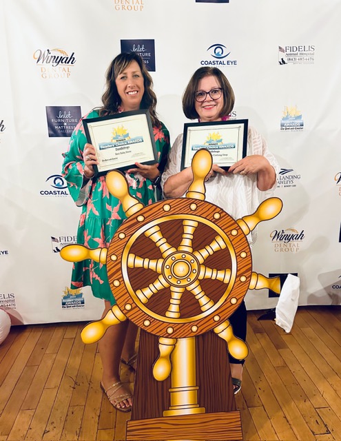Two women holding certificates stand behind a large ship's wheel prop on a wooden floor, smiling at the camera.
