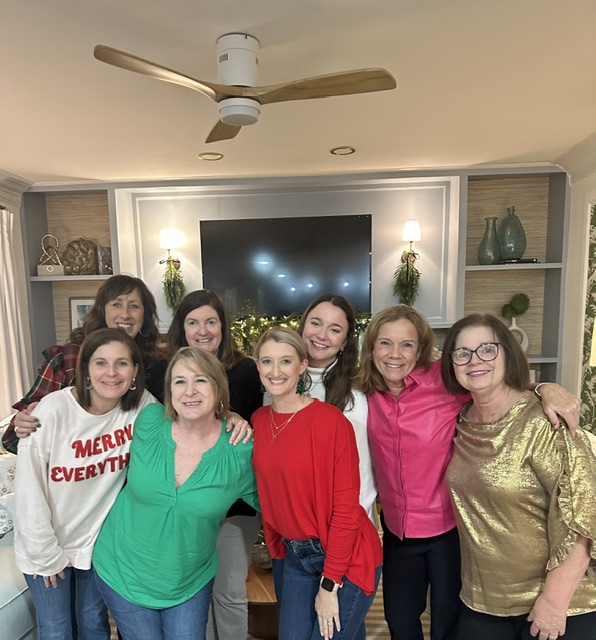 Group of seven women and girls smiling indoors, standing in front of a large TV, with shelves and decorative items behind them.