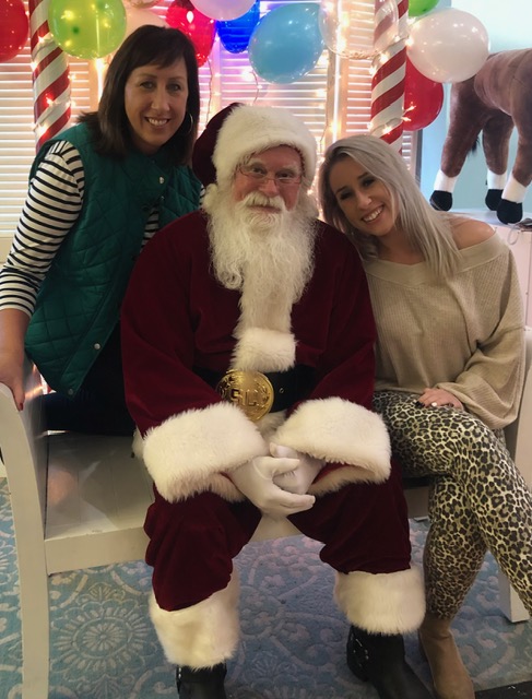 Two women and a man dressed as Santa Claus pose together indoors with holiday decorations and balloons in the background.
