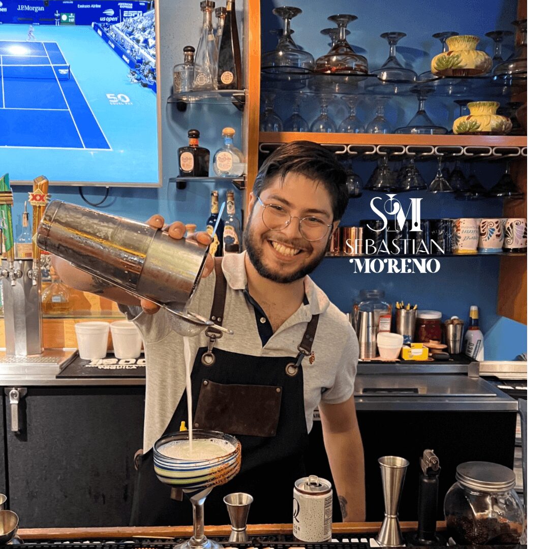 Bartender smiling, pouring drink into a glass, bar with bottles and glasses in background, tennis match on TV screen, person wearing apron