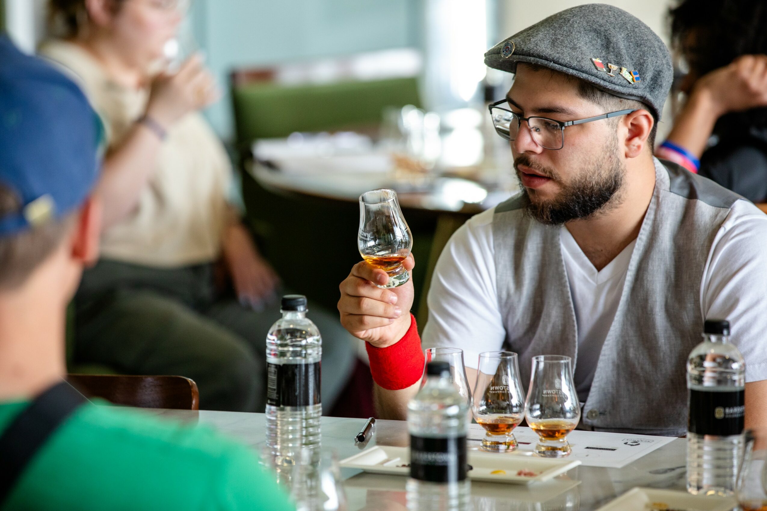 Man with glasses and a cap holding a glass of whiskey at a table with bottles and glasses, in a social setting.