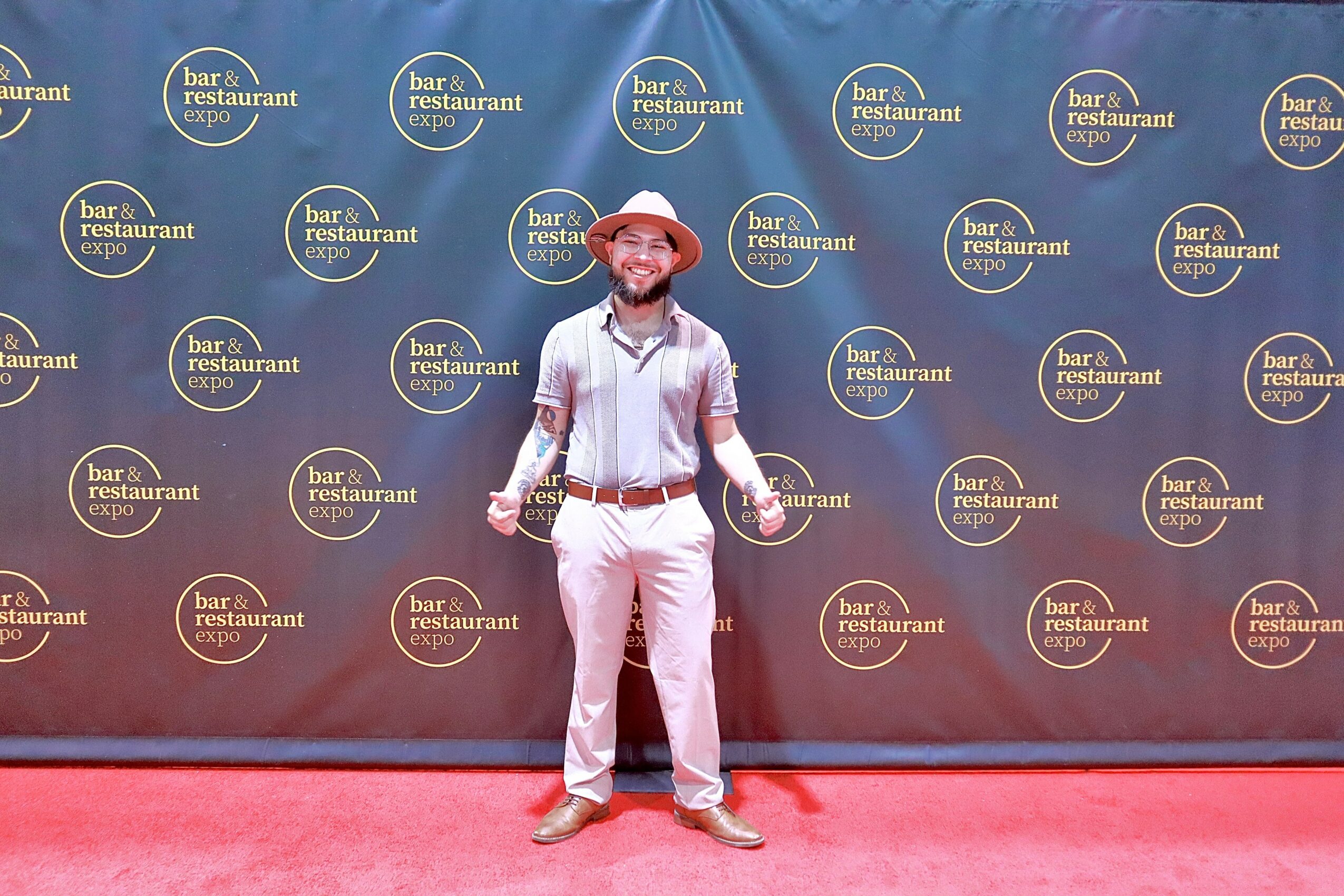Person standing on red carpet in front of a backdrop with repeated logo, smiling, wearing a hat, white shirt, and light-colored pants.
