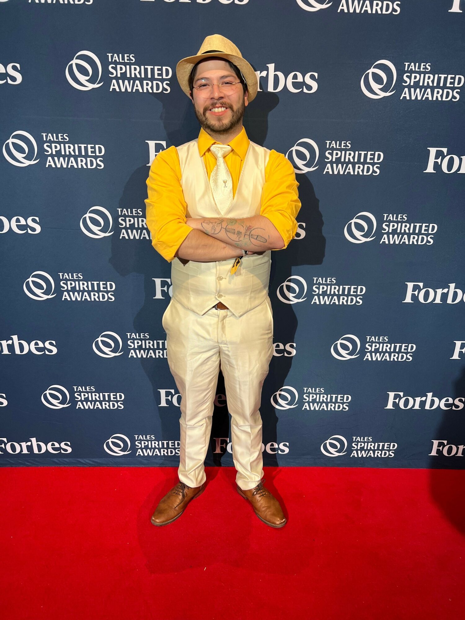 Person standing on red carpet in front of blue backdrop with logos, wearing yellow jacket and cream pants, smiling.