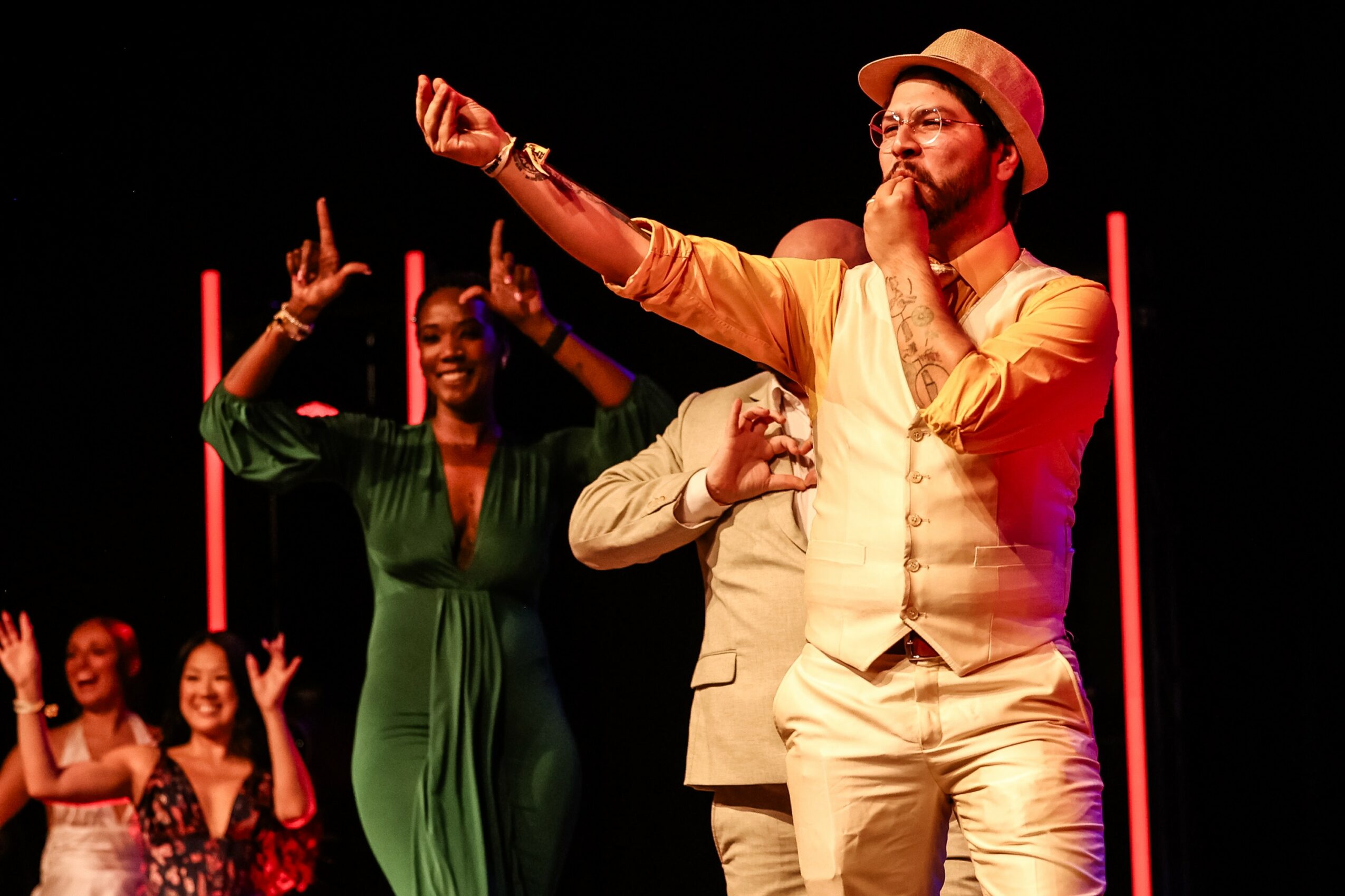 Group of people on stage, one man in a beige suit and hat taking a selfie, others smiling and raising hands, dark background
