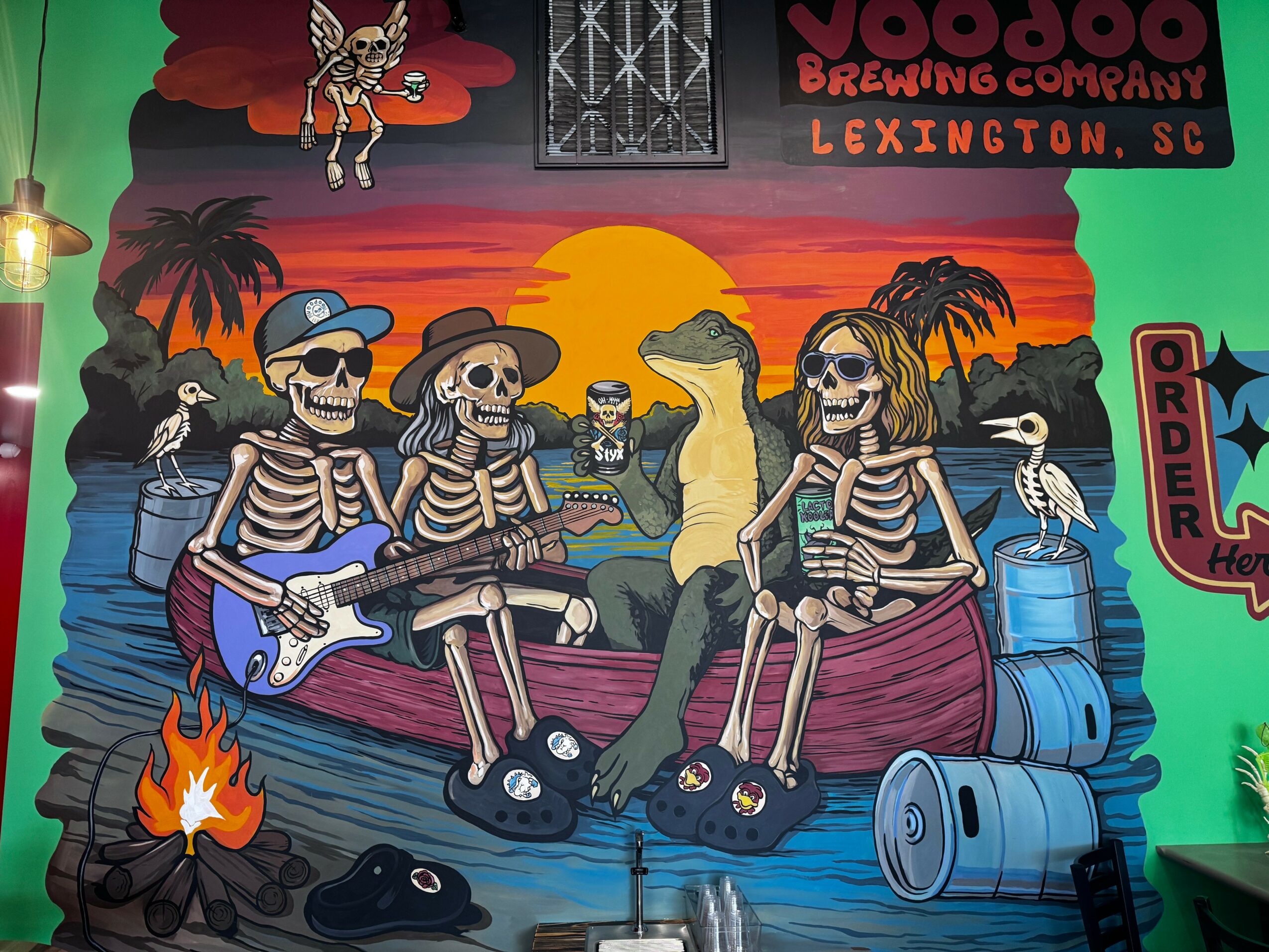 Colorful mural of skeletons in a boat with a crocodile, sunset, palm trees, and barrels, promoting Lexington, SC brewery.