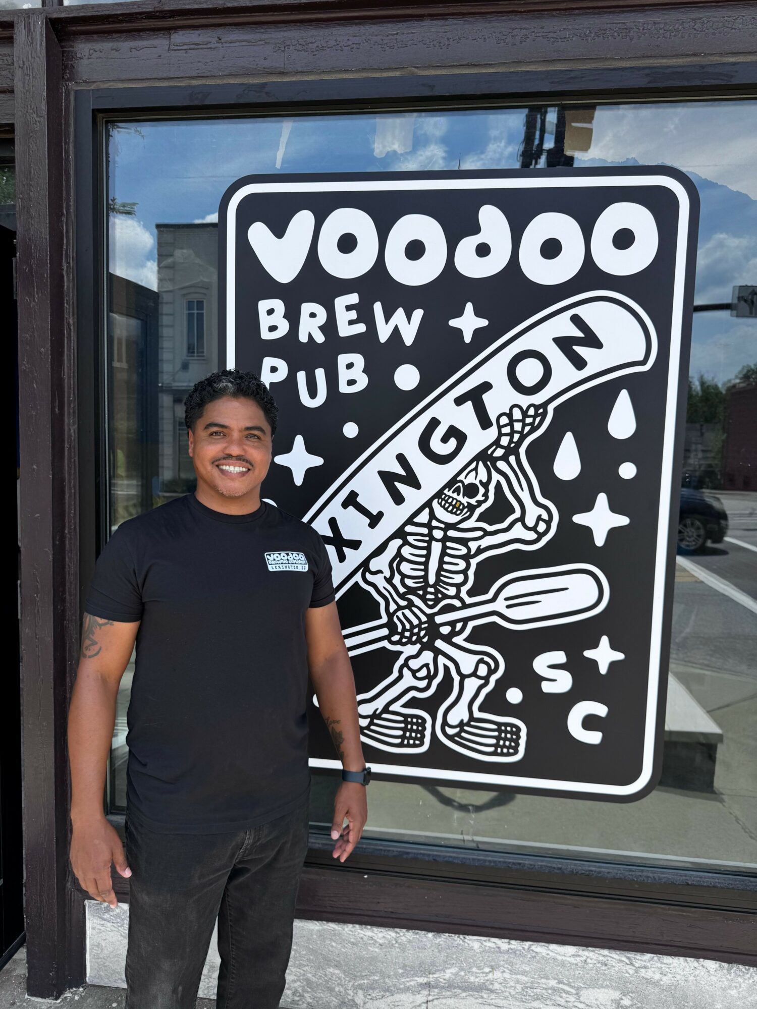 Person standing outside a window with a sign for Woods Brewpub and Kitchen, smiling, wearing a black shirt and pants.